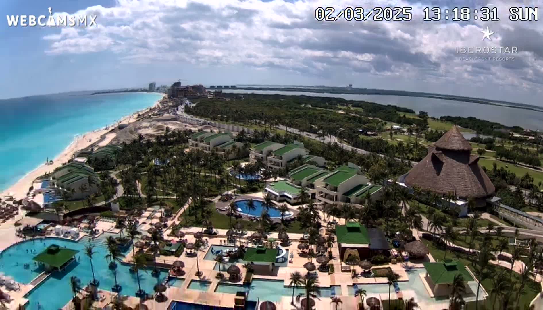 An aerial view shows a resort complex with multiple pools and buildings situated on a beach next to a calm turquoise ocean under partly cloudy skies.