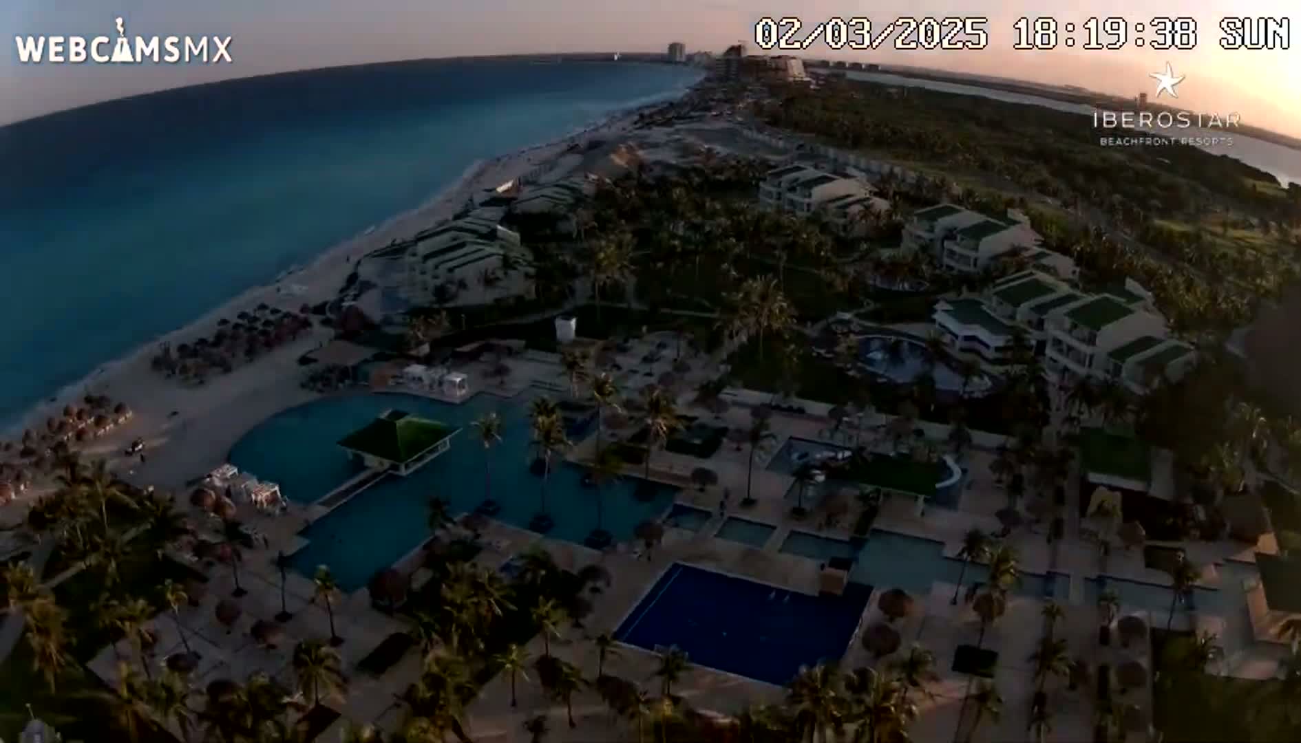 An aerial view shows a beachfront resort at sunset, featuring multiple pools, palm trees, and buildings along a sandy beach with calm, turquoise water.