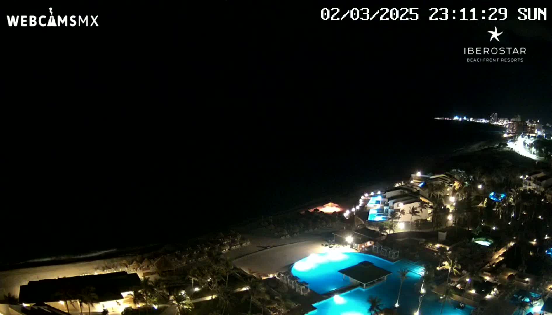 A nighttime aerial view shows a resort complex with illuminated pools and buildings along a dark ocean beach under a clear, dark sky.
