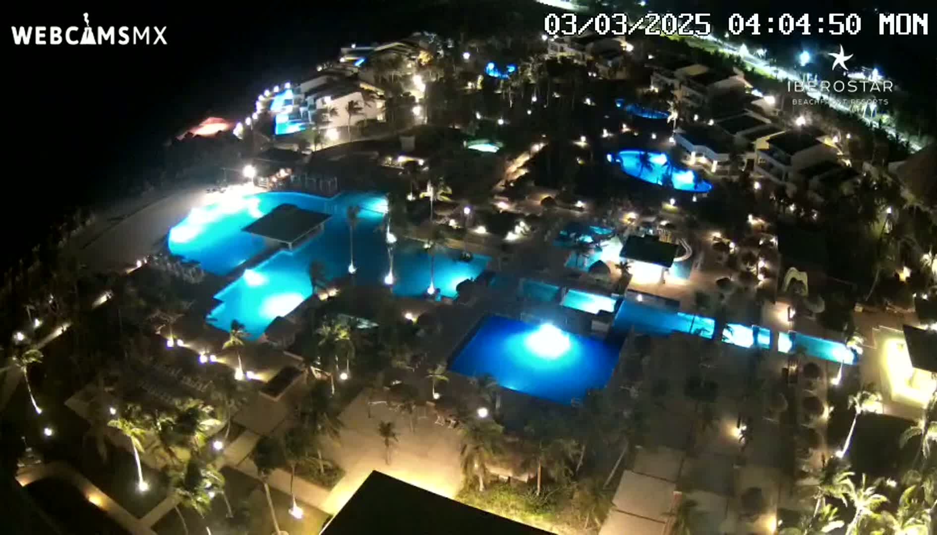 A nighttime aerial view of a resort complex with numerous illuminated swimming pools surrounded by palm trees and buildings.