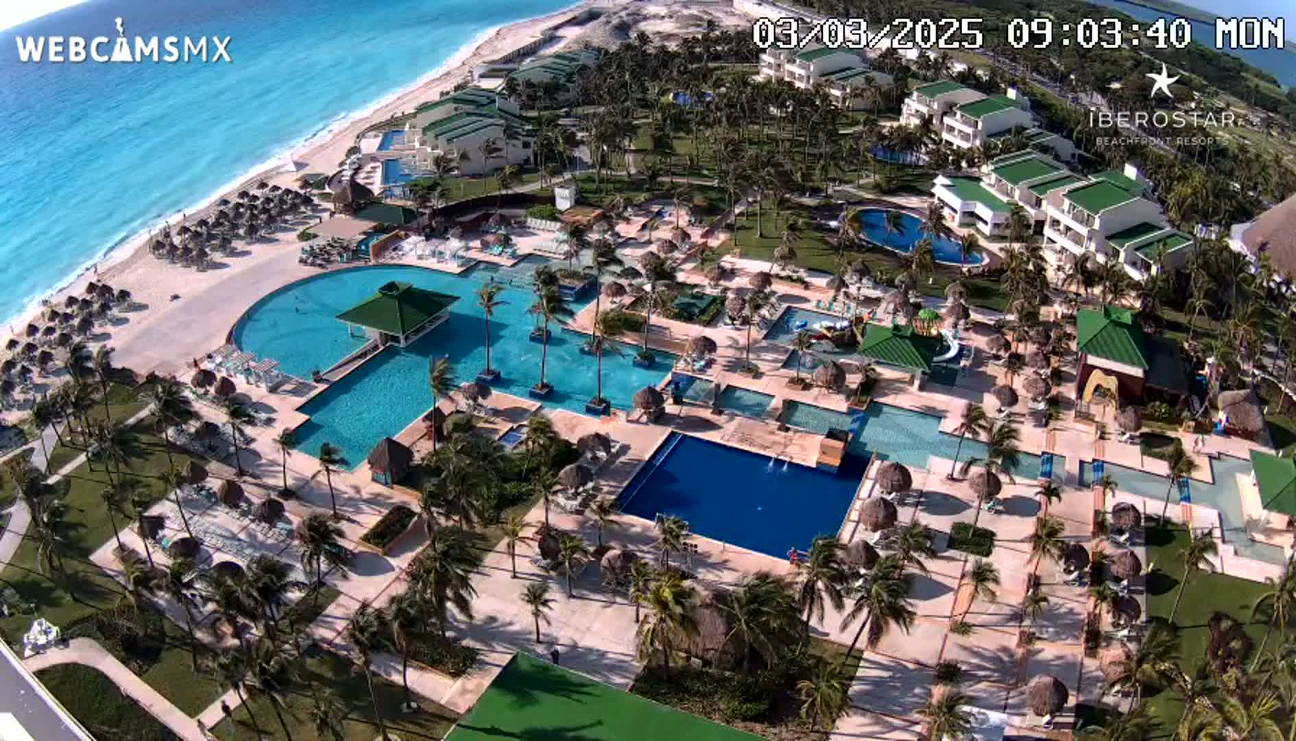 An aerial view shows a resort complex with numerous pools, palm trees, and buildings situated next to a beach with turquoise water under sunny conditions.