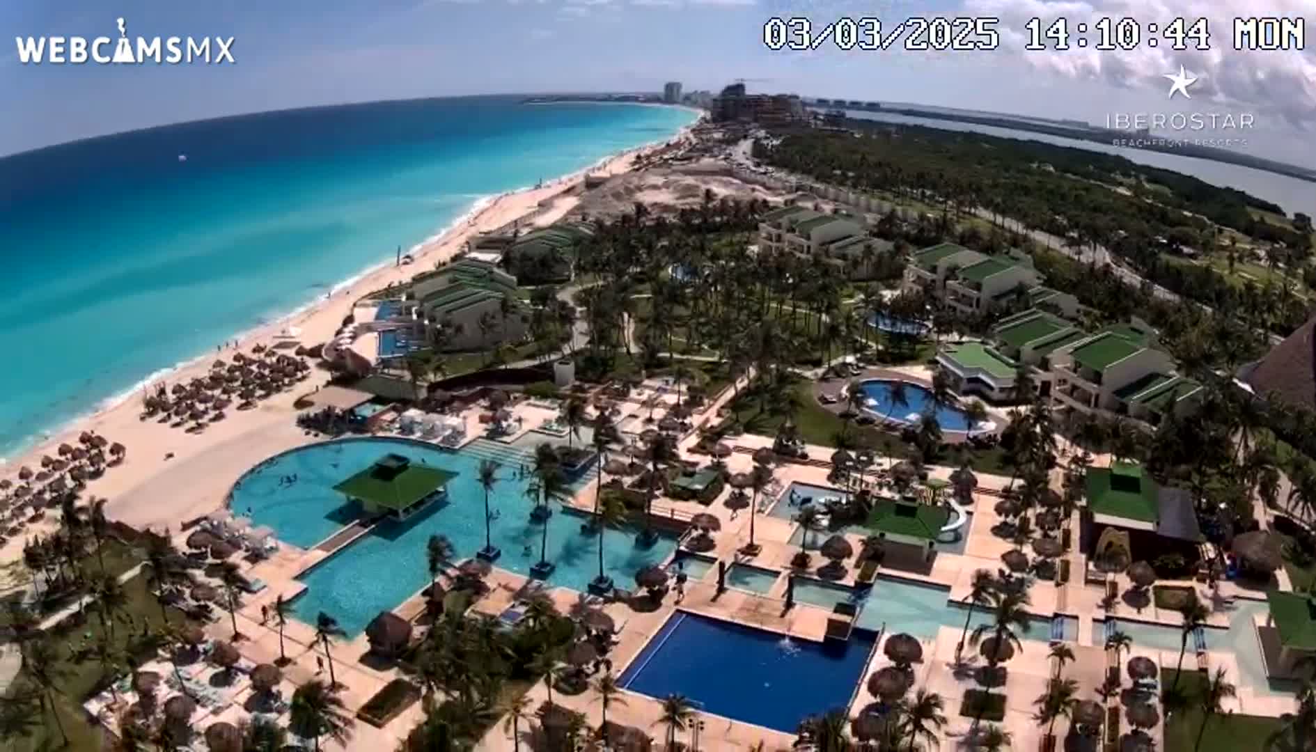 An aerial view shows a resort with numerous pools and a beach on a sunny day with clear turquoise water.