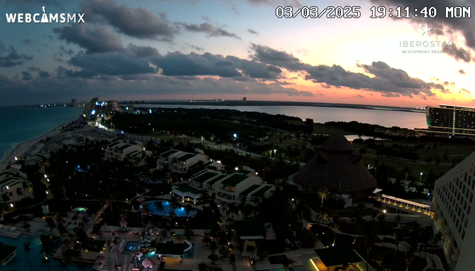 An aerial, nighttime view of a resort town on a coast at sunset, with partly cloudy skies and calm water.