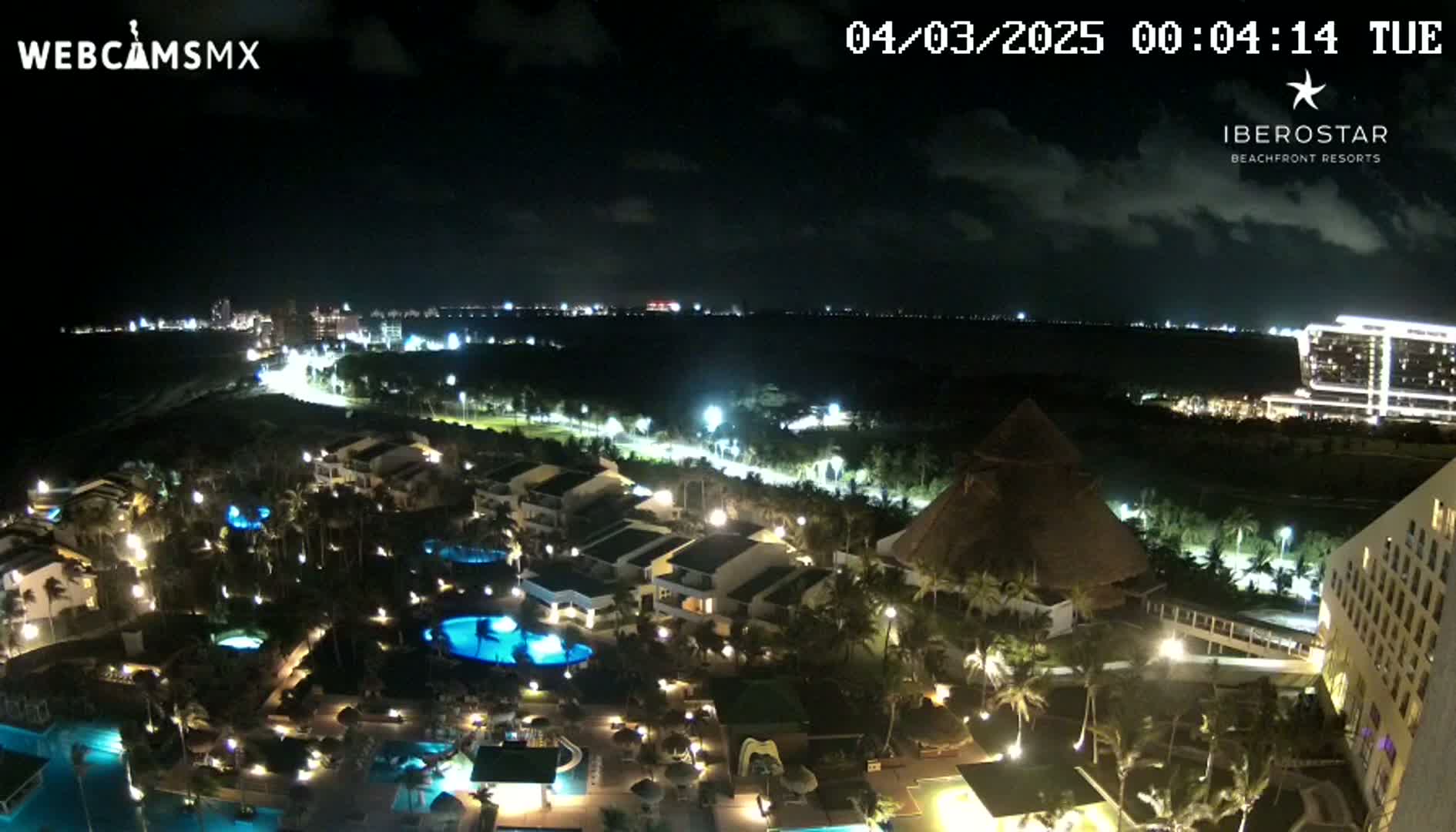 A nighttime aerial view of a resort area showing lit buildings, pools, and palm trees, with a calm ocean in the distance under a partly cloudy sky.
