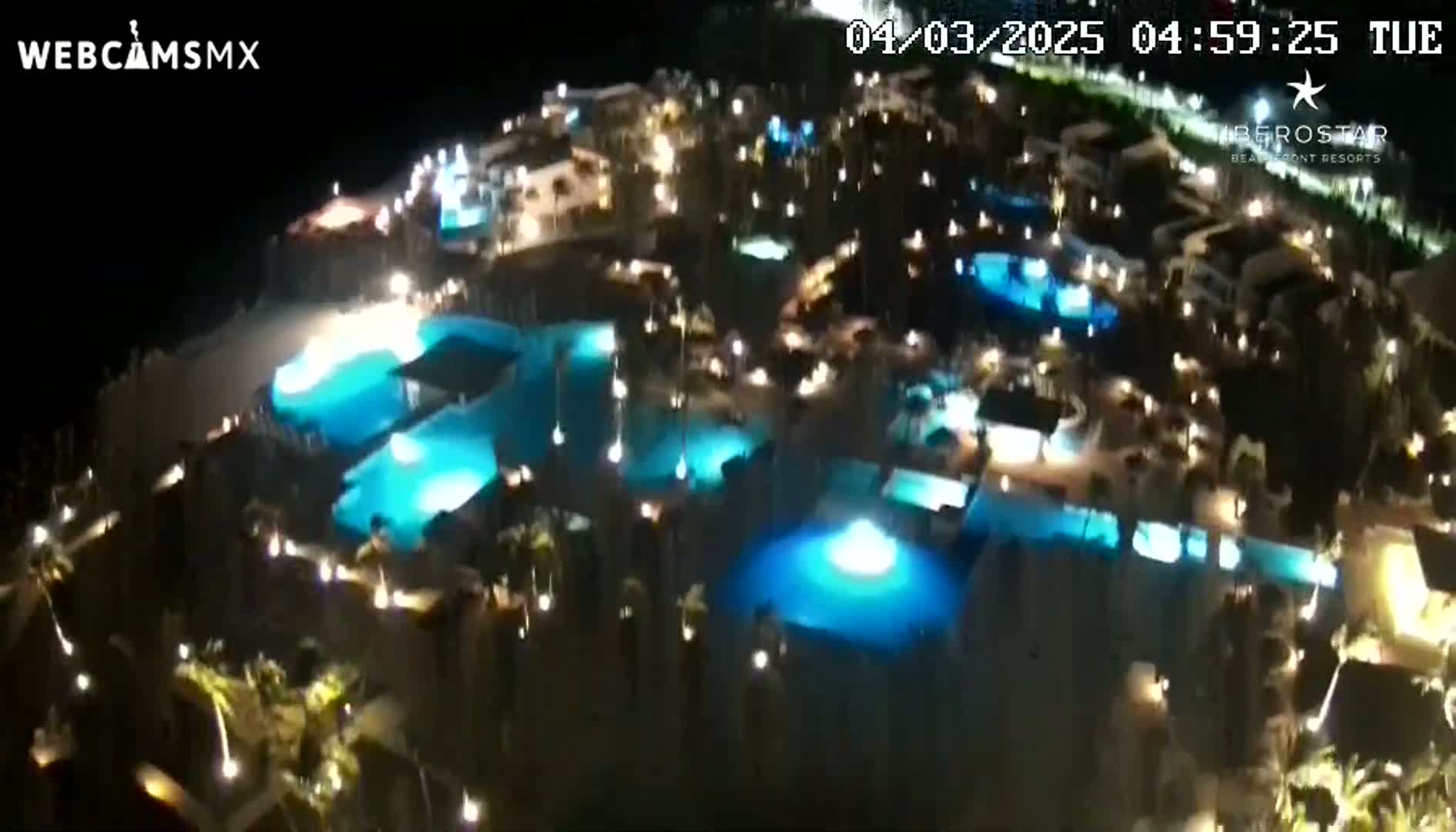 An aerial nighttime view shows numerous illuminated swimming pools surrounded by buildings and landscaping.