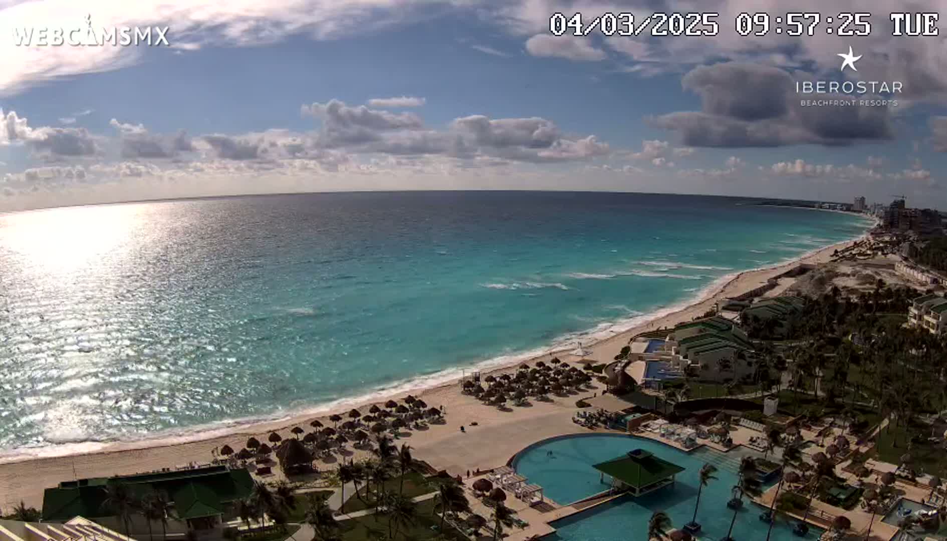 A mostly sunny day shows a long stretch of beach with turquoise water, a resort with multiple pools, and palm trees under a partly cloudy sky.
