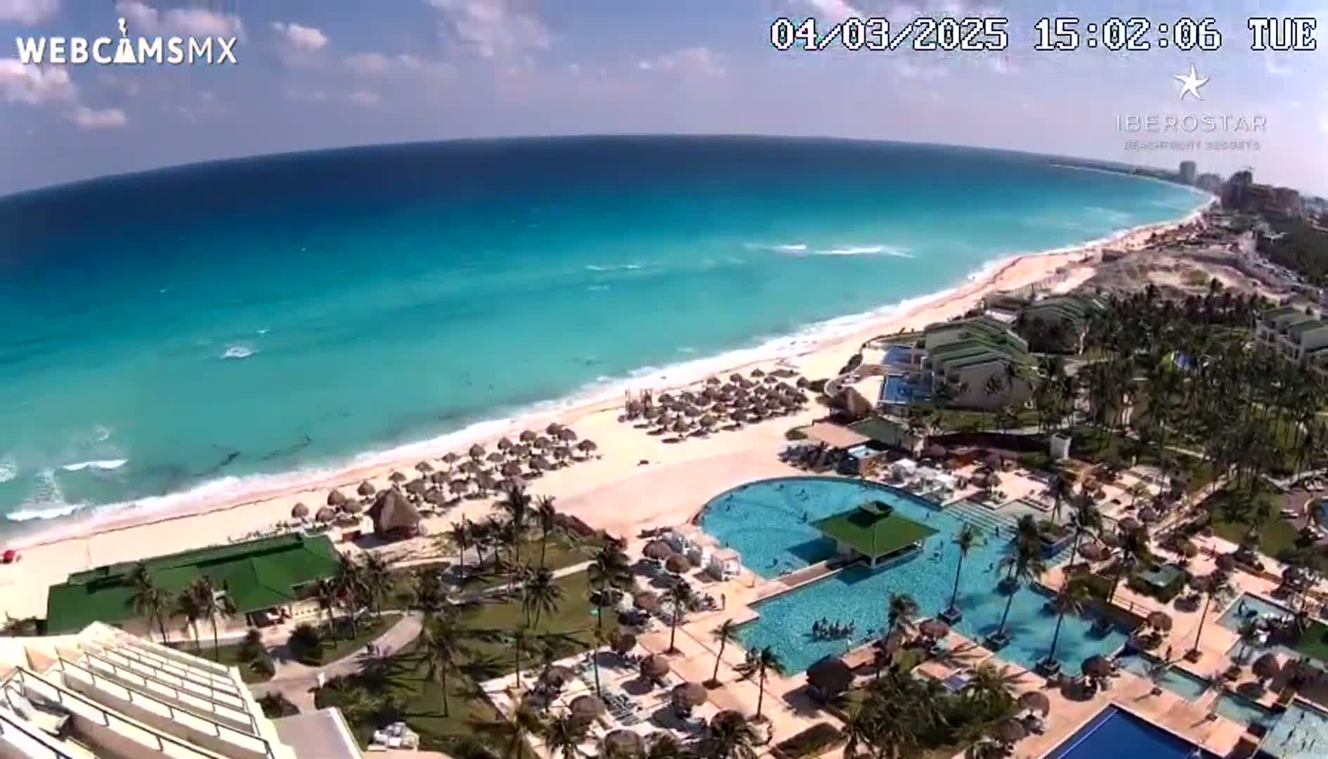 An aerial view shows a resort complex with multiple pools and a beach on a sunny day with turquoise water and small waves.