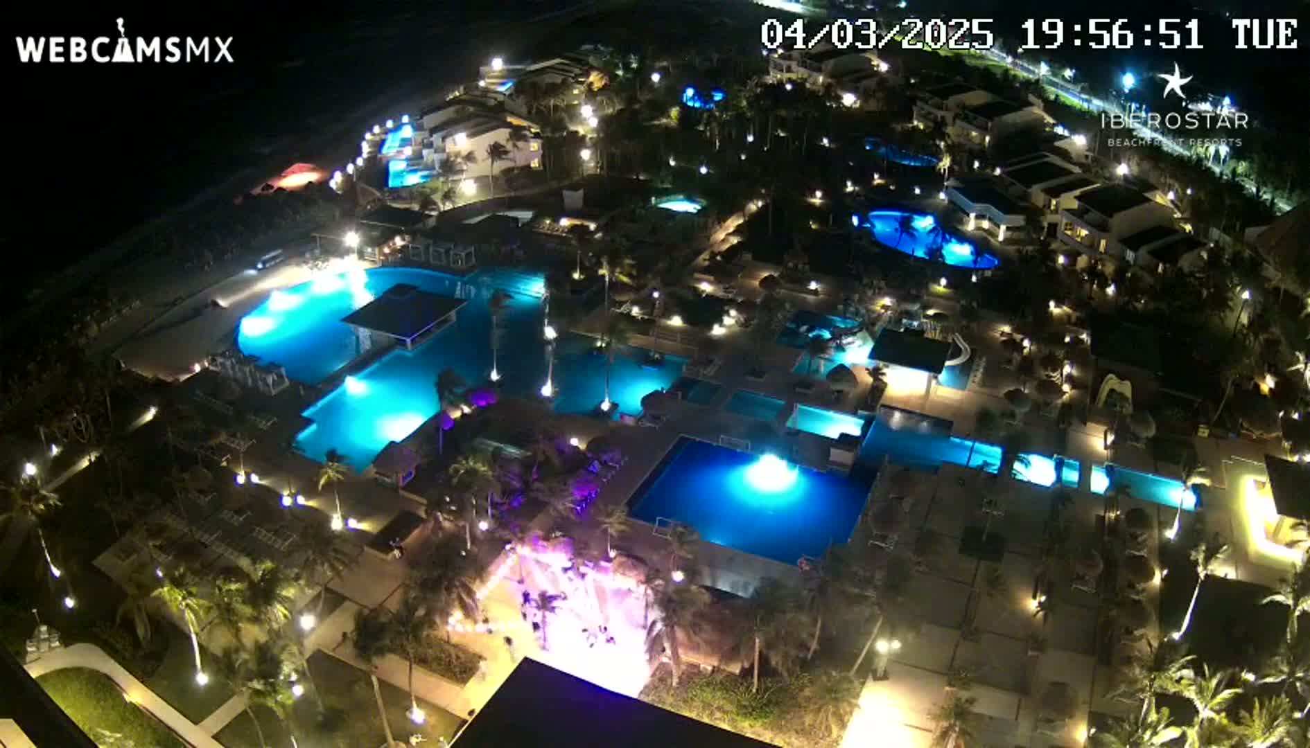 An aerial nighttime view shows a resort complex with numerous illuminated pools and surrounding buildings, under clear skies.