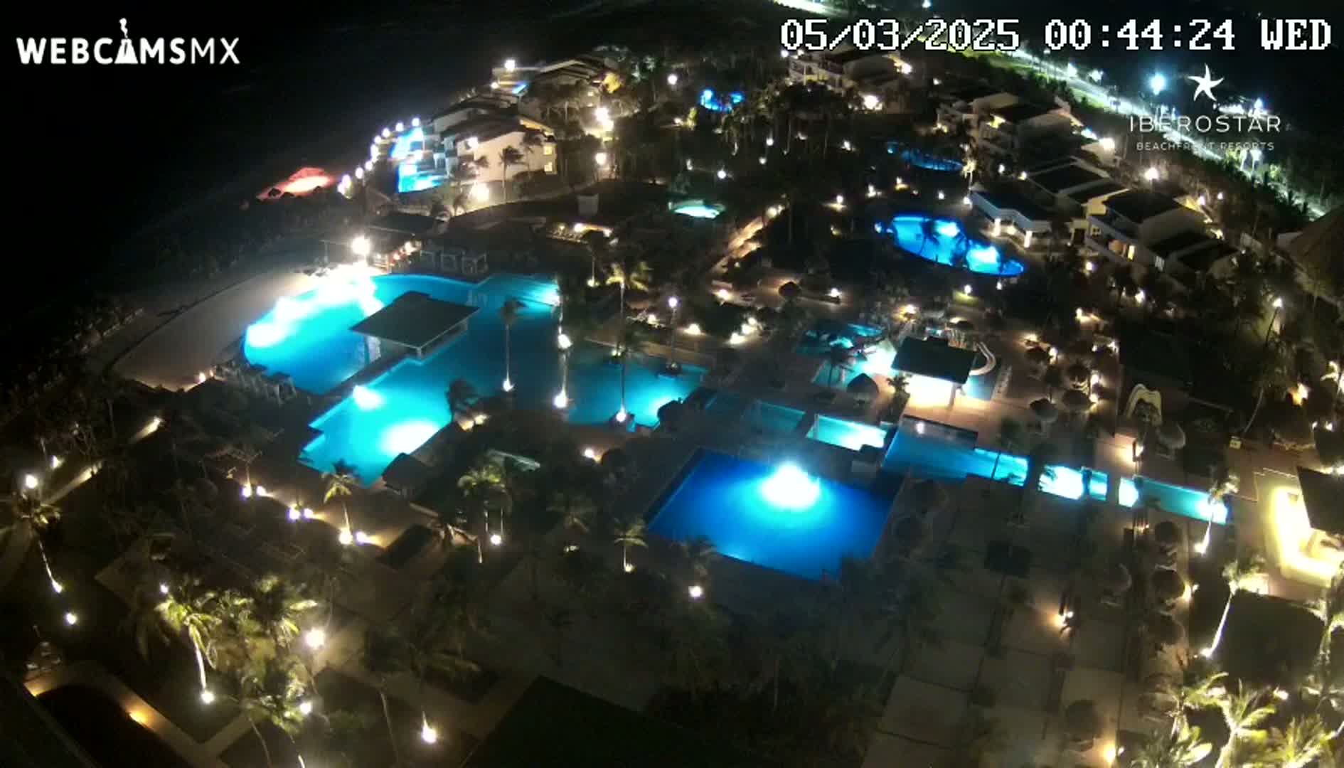 An aerial nighttime view shows a resort complex with numerous illuminated swimming pools surrounded by palm trees and buildings.