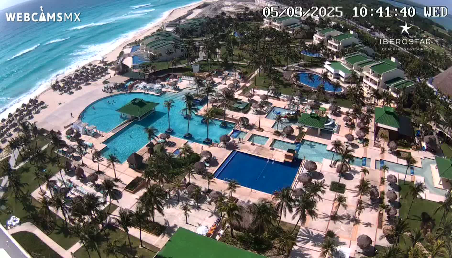 An aerial view of a resort with multiple pools and palm trees surrounding buildings, next to a beach with turquoise water and small waves under a sunny sky.