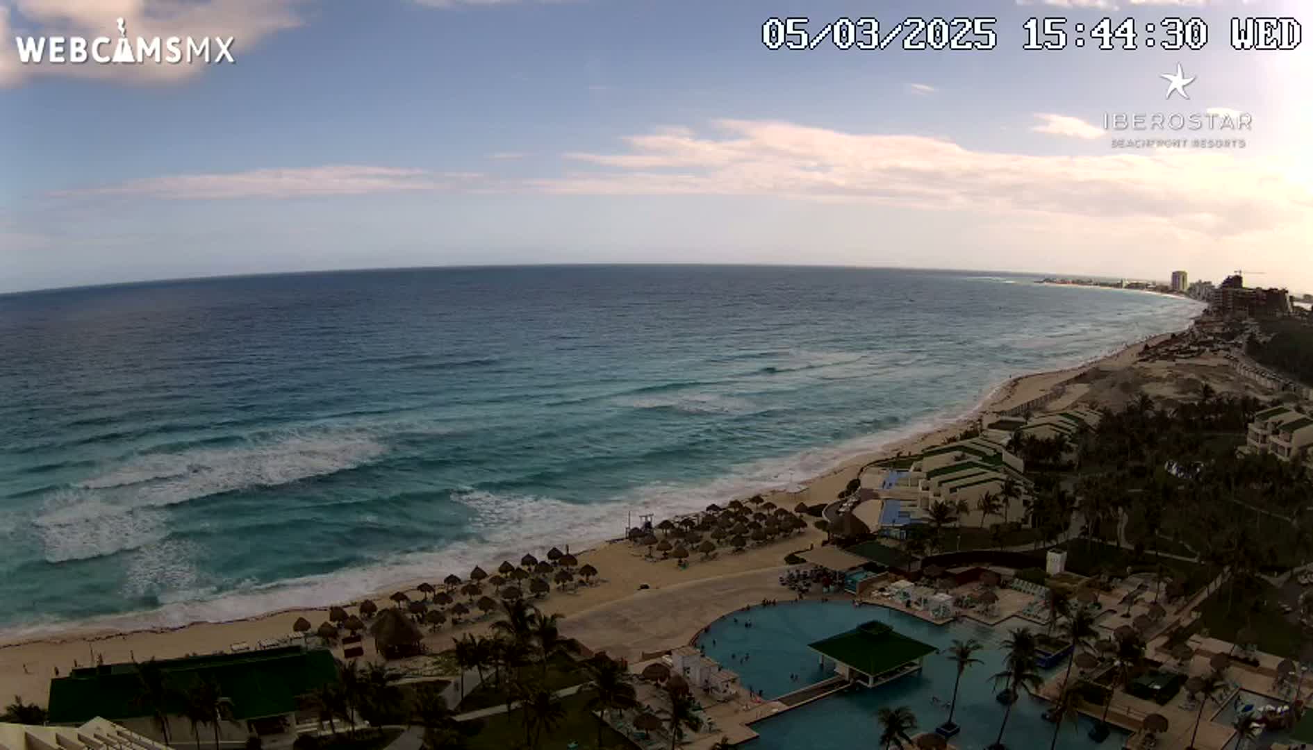 A mostly sunny, slightly hazy beach scene shows a resort with swimming pools nestled on a sandy shoreline, where gentle waves lap the turquoise ocean.