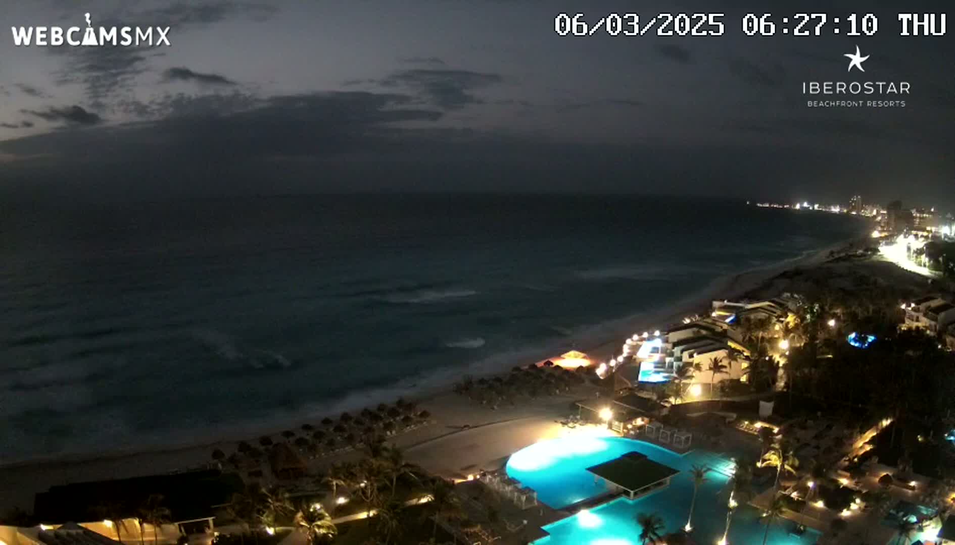 A nighttime, coastal resort scene shows a dark ocean with small waves under a cloudy sky, with the resort's illuminated buildings, pools, and beach umbrellas visible along the shoreline.