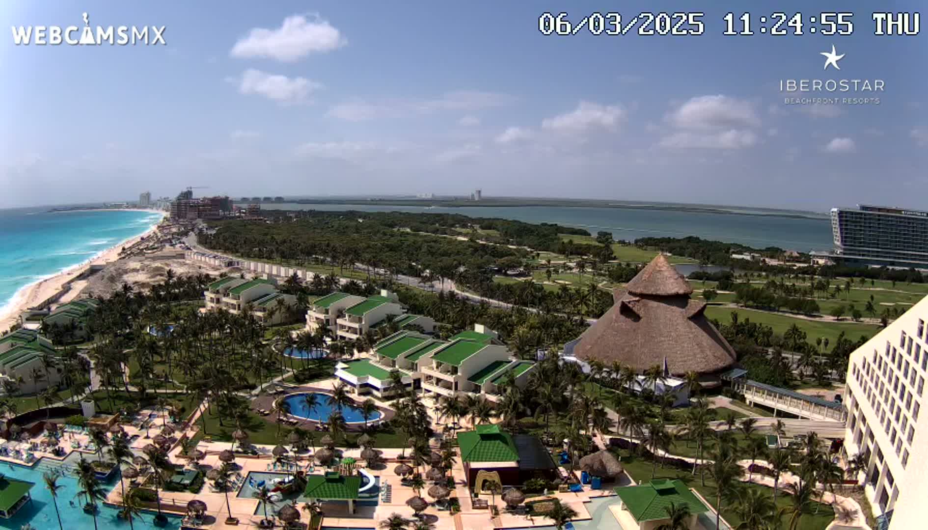 An aerial view of a resort complex on a sunny day, featuring multiple buildings, swimming pools, lush landscaping, and a beach with turquoise water in the background.
