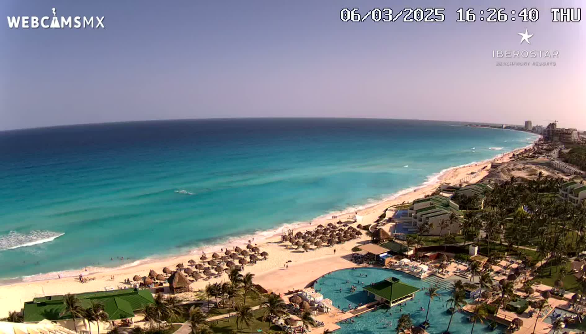 A clear, sunny day shows a long stretch of beach with turquoise water, lined with resorts, palm trees, and beach umbrellas.