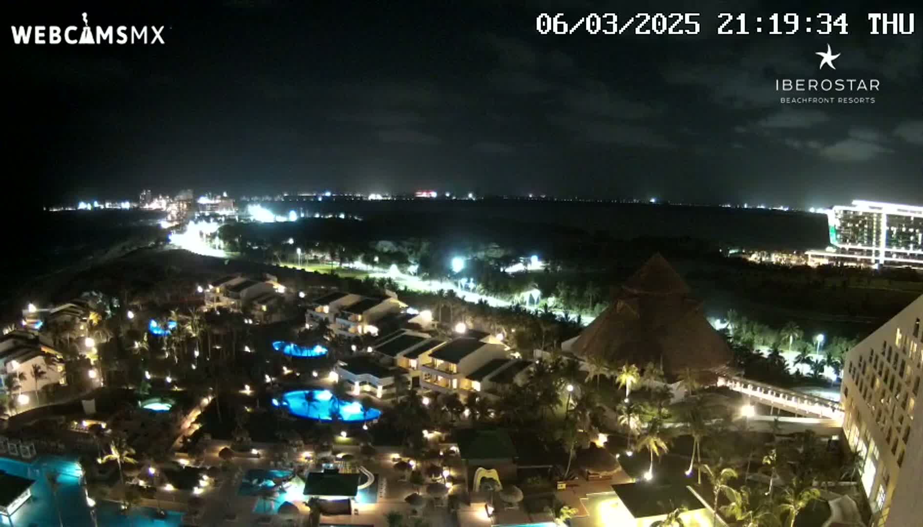 A nighttime aerial view of a resort complex with pools and palm trees, illuminated buildings, and a body of water in the background under a clear, dark sky.