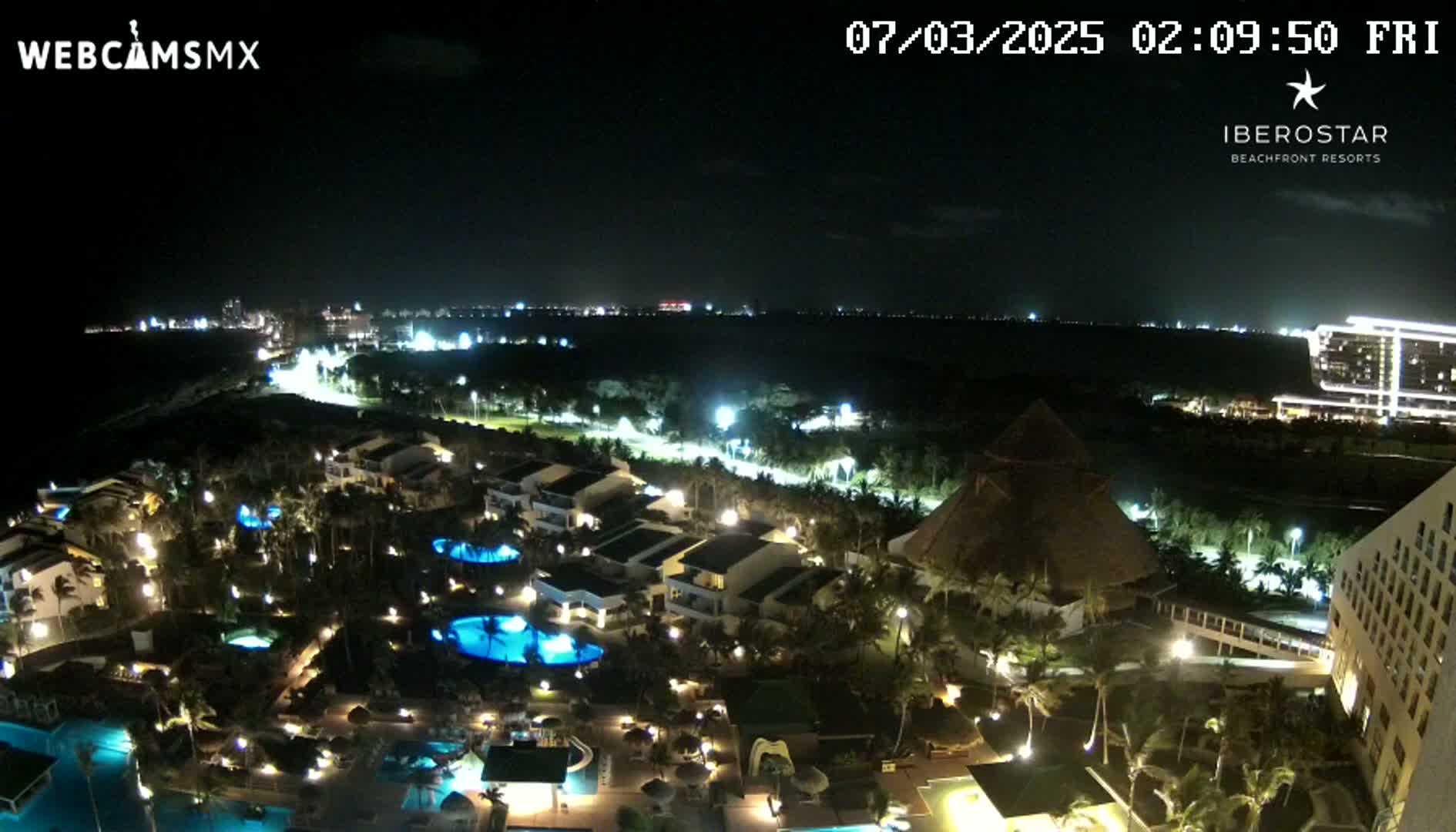 A nighttime aerial view of a resort complex with illuminated buildings, pools, and landscaping, overlooking a dark body of water and a distant city skyline under a clear sky.