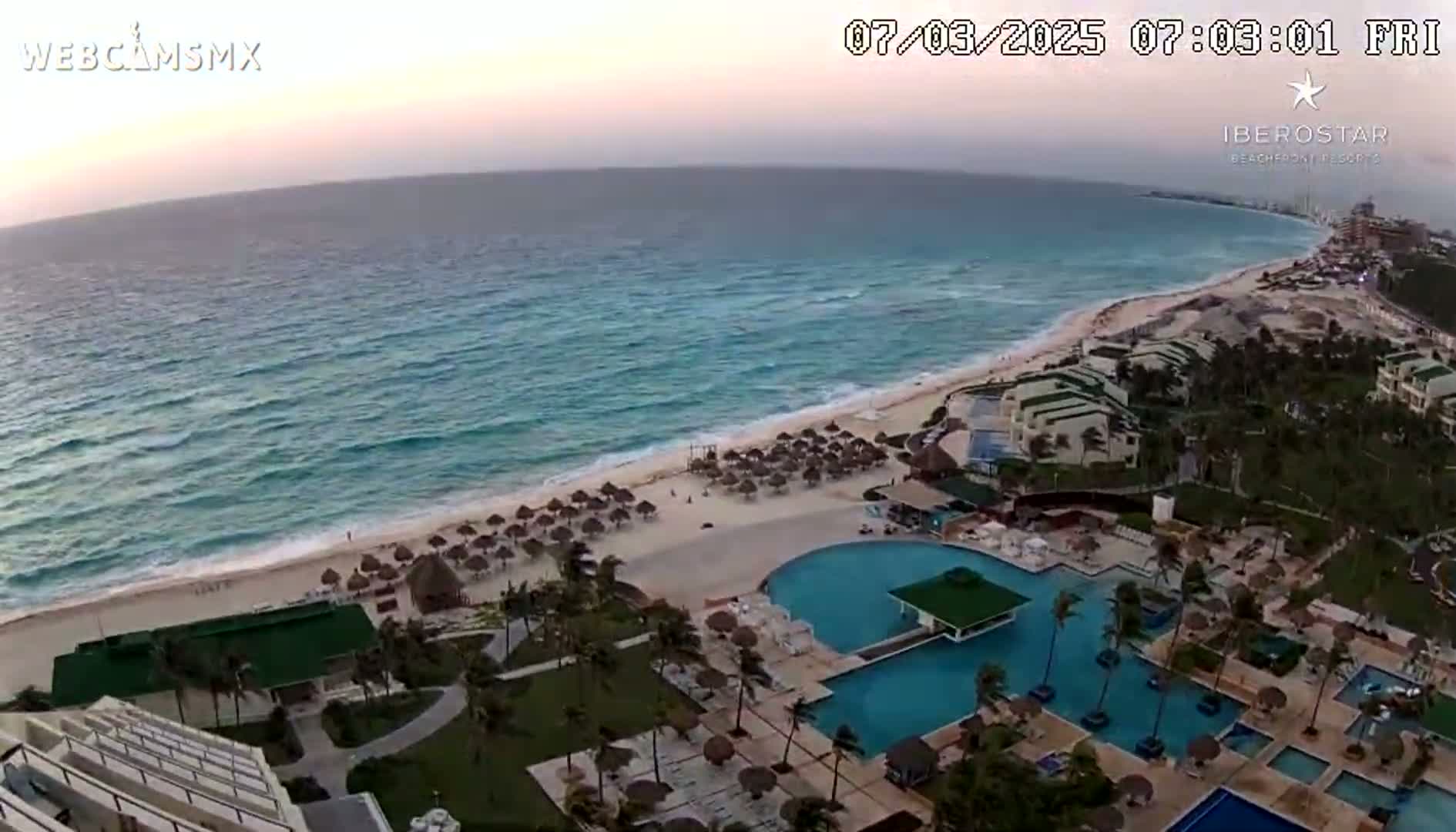 An aerial view shows a resort with multiple pools and a beach at sunset, with moderate waves in the turquoise ocean.