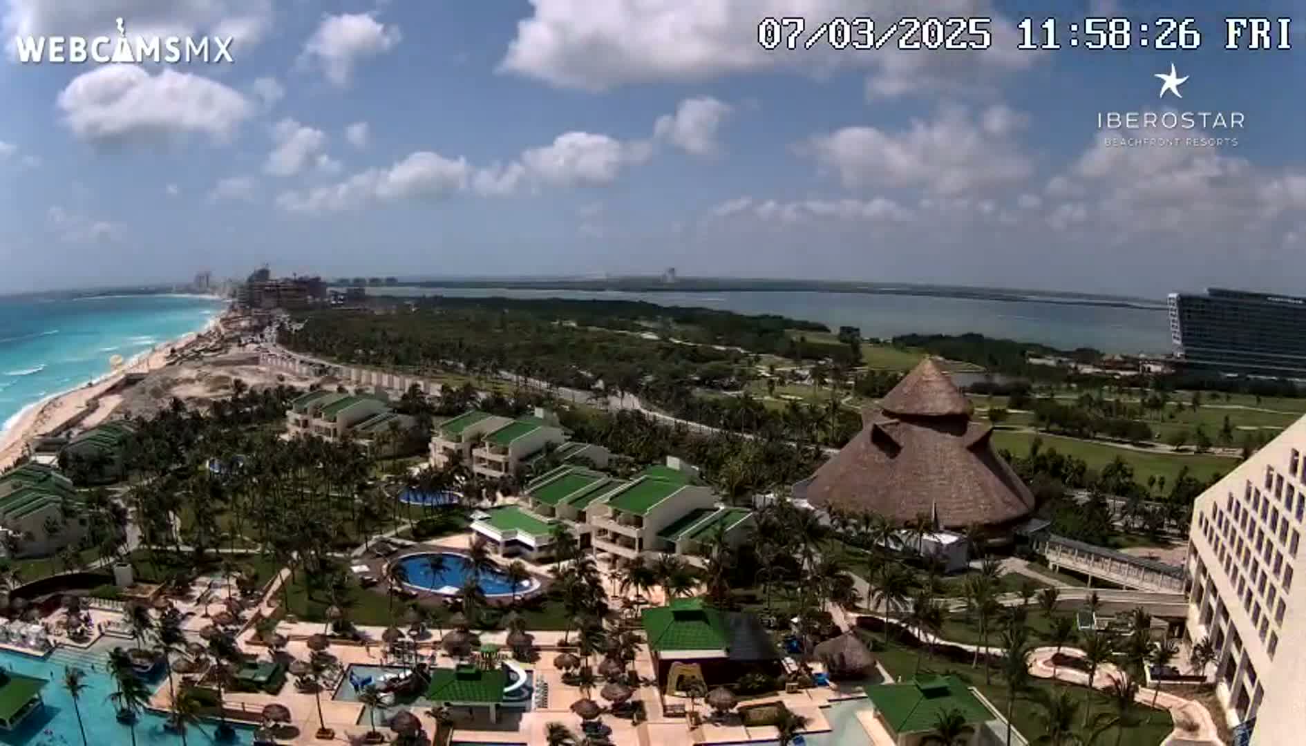 An aerial view of a resort complex on a partly cloudy day, showing buildings, pools, landscaping, and a beach with turquoise water in the background.