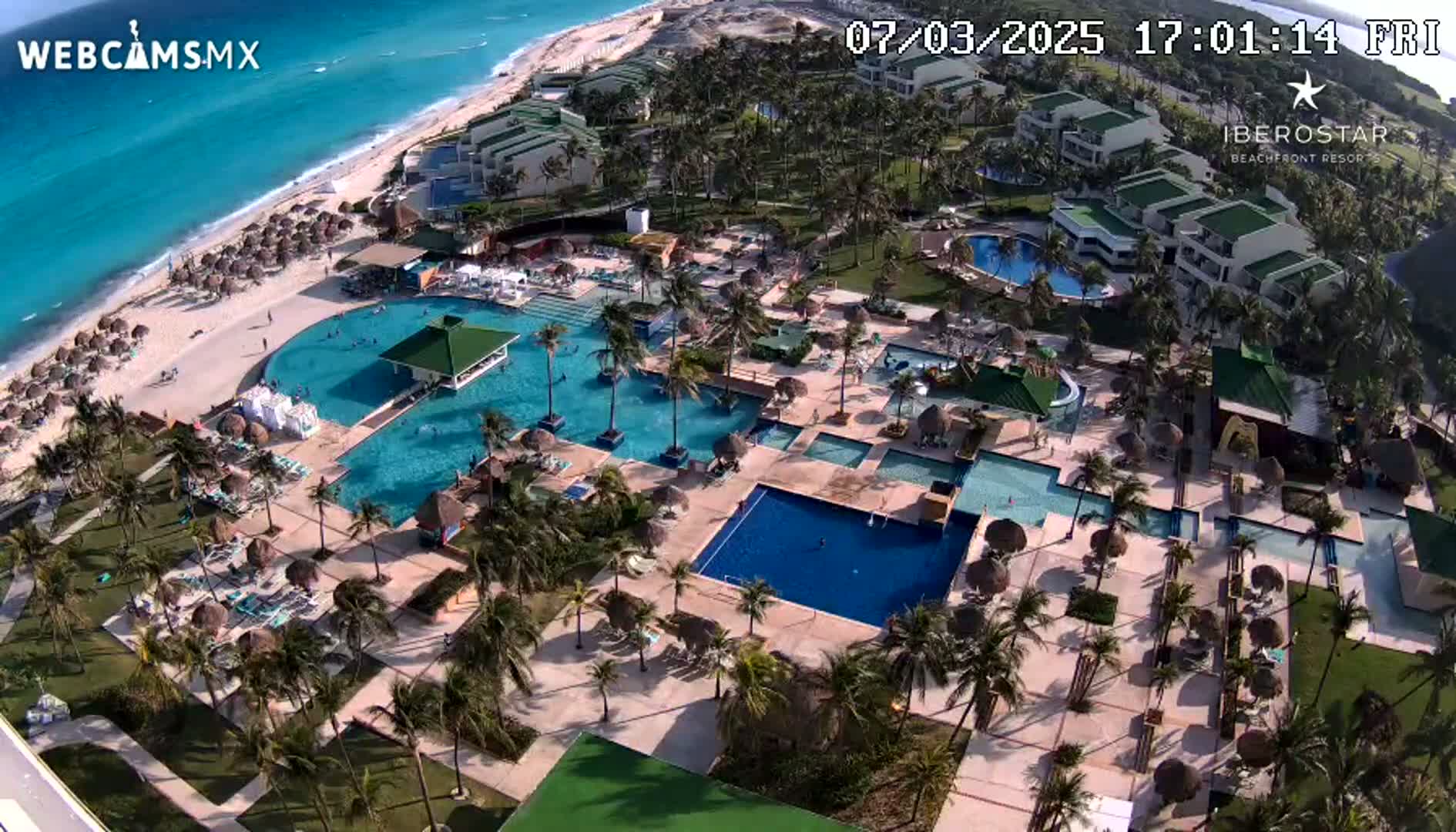 An aerial view shows a resort with multiple pools, palm trees, and a beach with clear turquoise water under sunny skies.