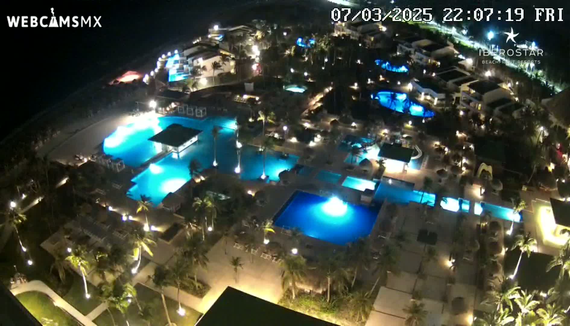 An illuminated resort at night, featuring numerous pools and palm trees surrounding low-rise buildings.