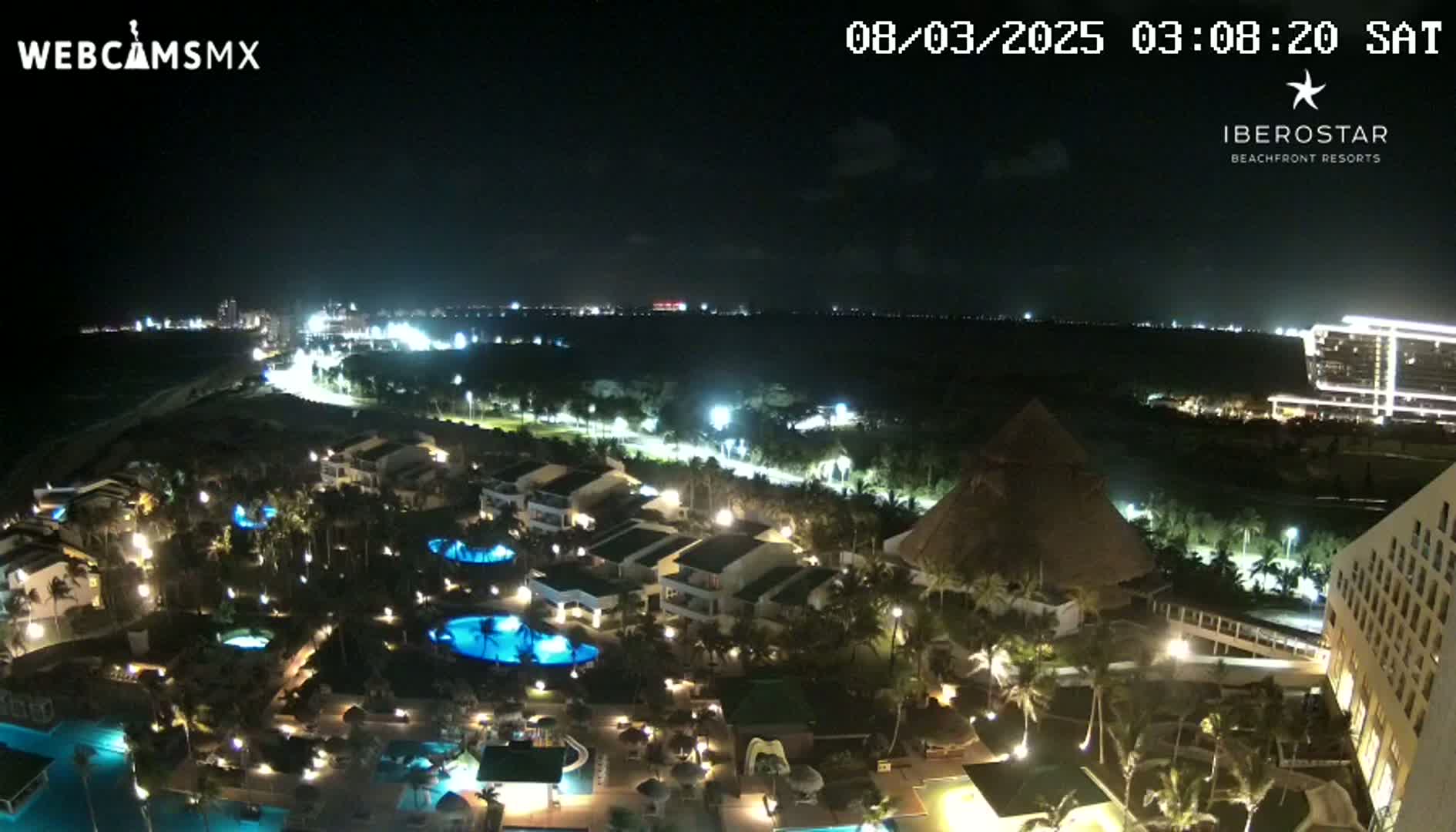 A nighttime aerial view of a resort complex with multiple pools, illuminated buildings, and palm trees, overlooking a coastline under a clear sky.