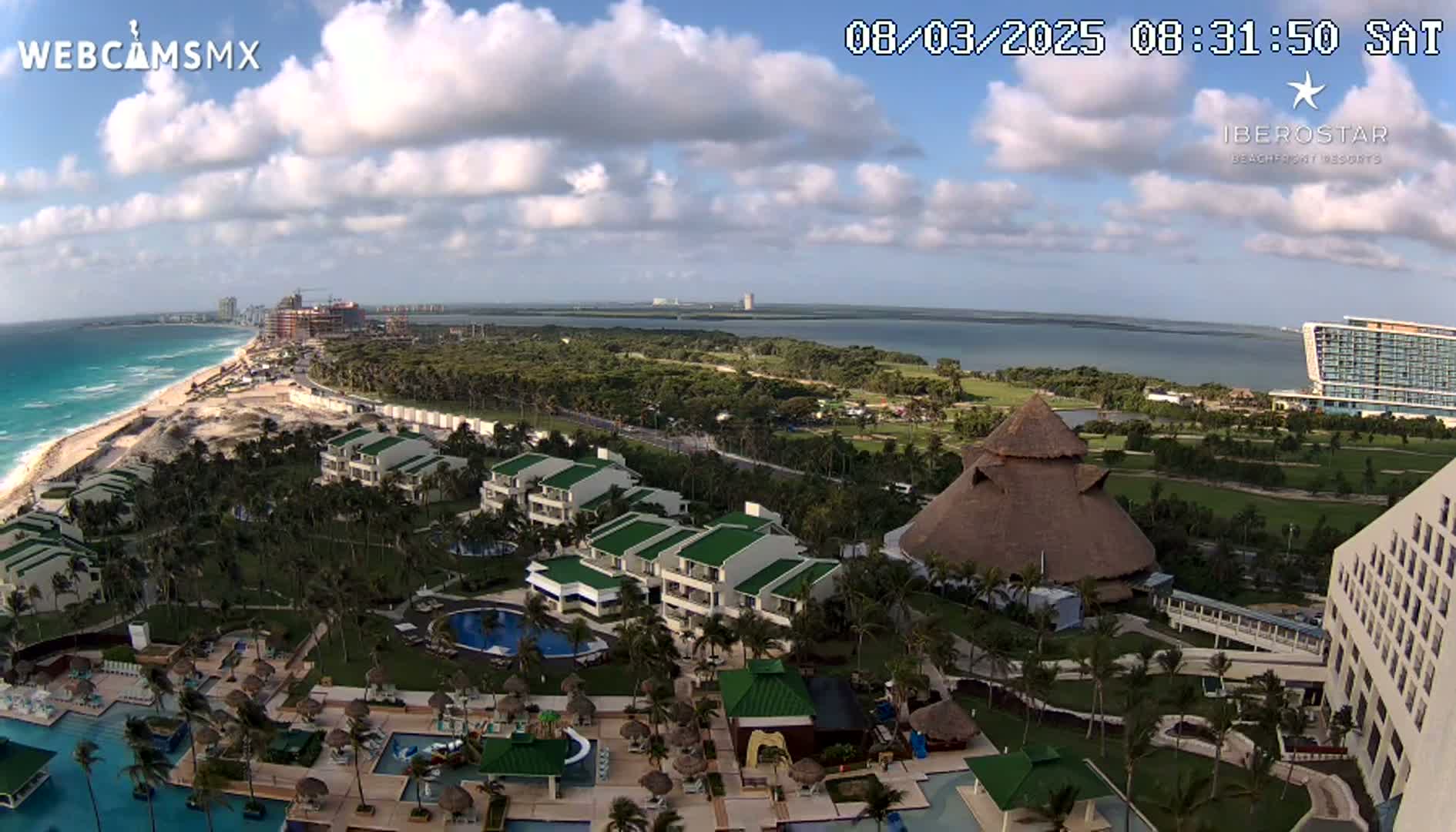 An aerial view of a resort complex on a partly cloudy day, showing turquoise ocean waves, a beach, and various buildings including a large thatched-roof structure.