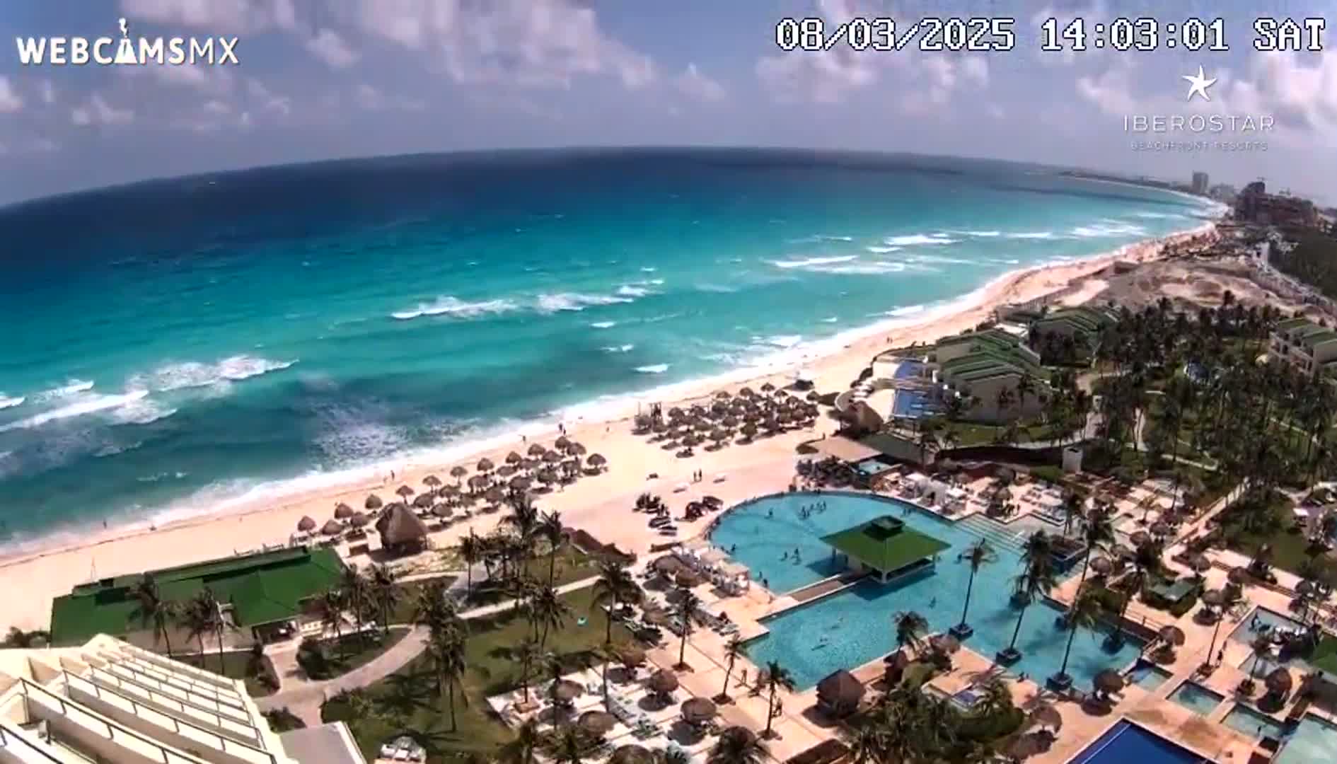 An aerial view shows a resort complex with multiple pools next to a beach with turquoise water and waves under a mostly sunny sky.