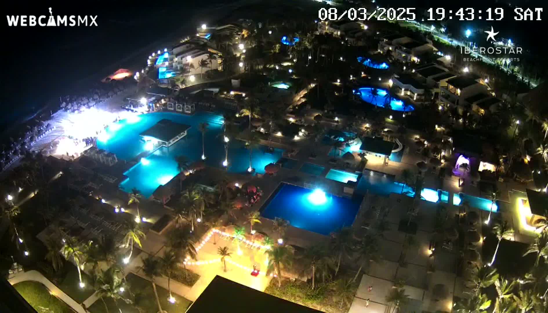 An illuminated resort at night, featuring numerous pools and palm trees, is seen from an elevated vantage point.