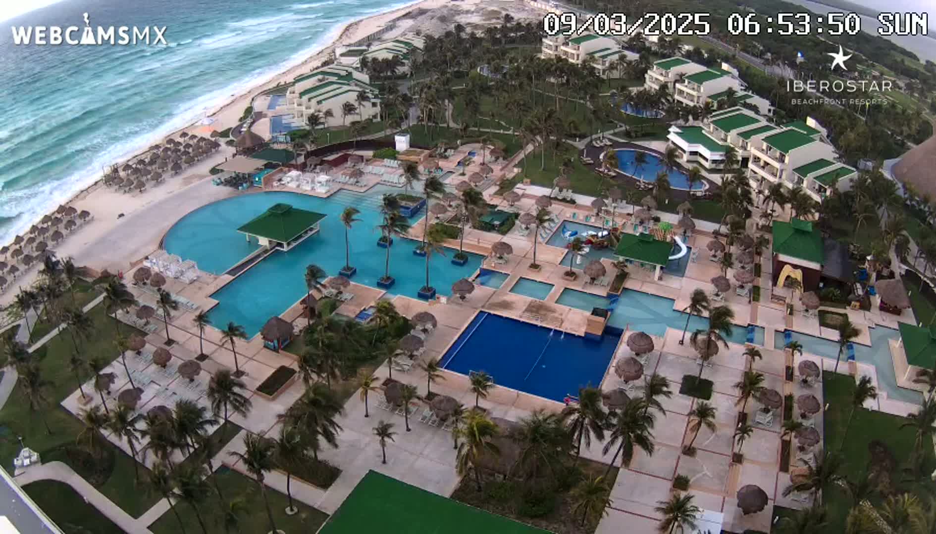An aerial view of a resort with multiple pools, beach umbrellas, and buildings on a sunny day with windy conditions near an ocean with waves.