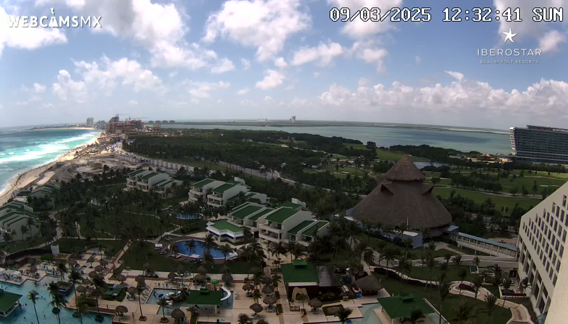 An aerial view of a resort complex on a partly cloudy day, showcasing buildings, swimming pools, landscaping, and a beach with ocean waves visible in the distance.