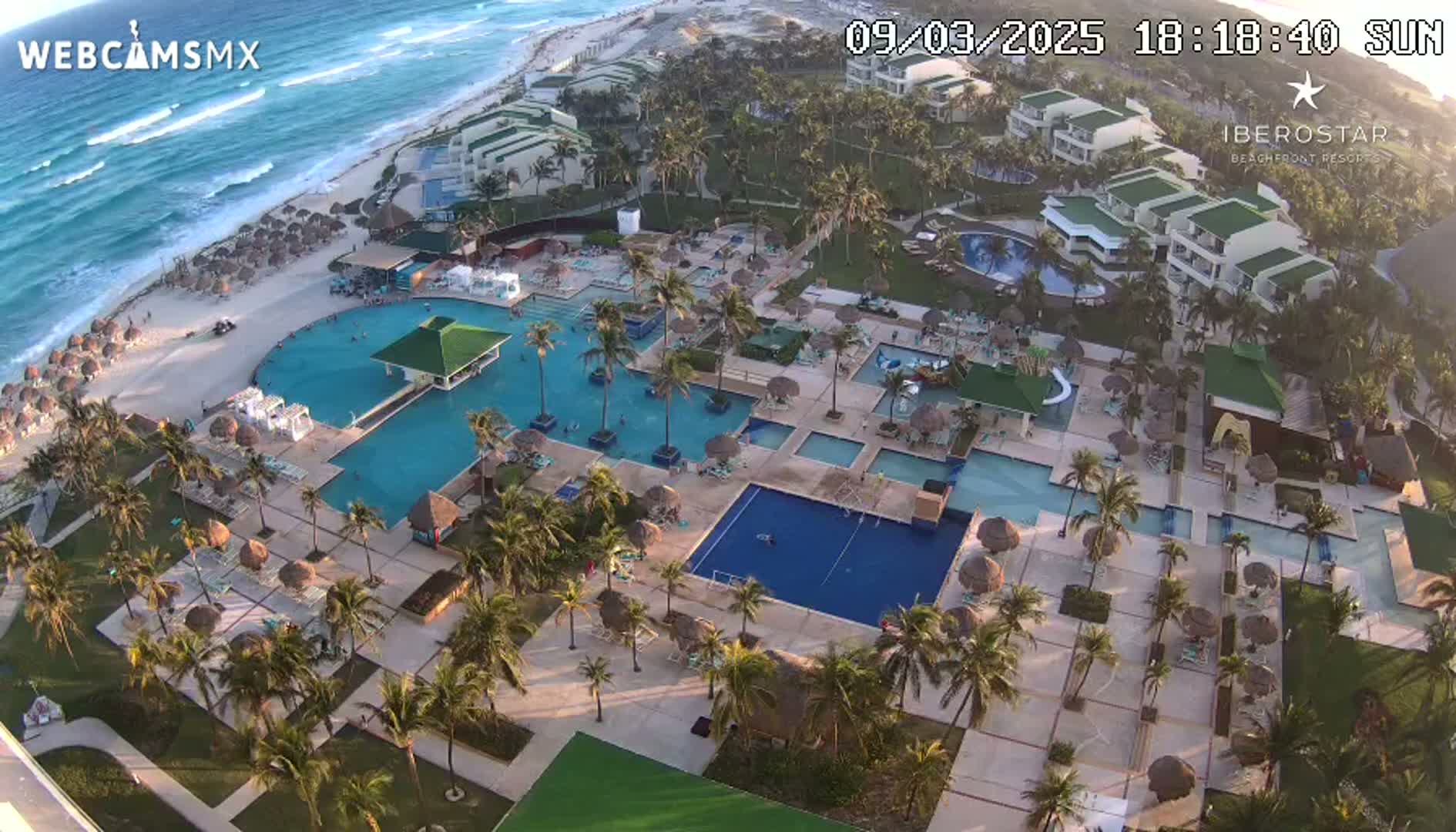 An aerial view of a resort on a sunny day shows multiple pools surrounded by palm trees and buildings, next to a beach with waves visible in the ocean.