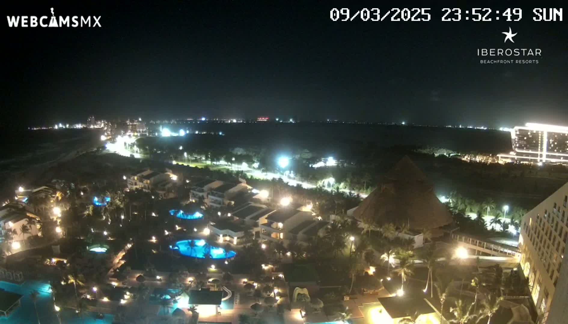 A nighttime aerial view of a resort complex with swimming pools and palm trees illuminated, overlooking a dark ocean and a city skyline in the distance under a clear sky.