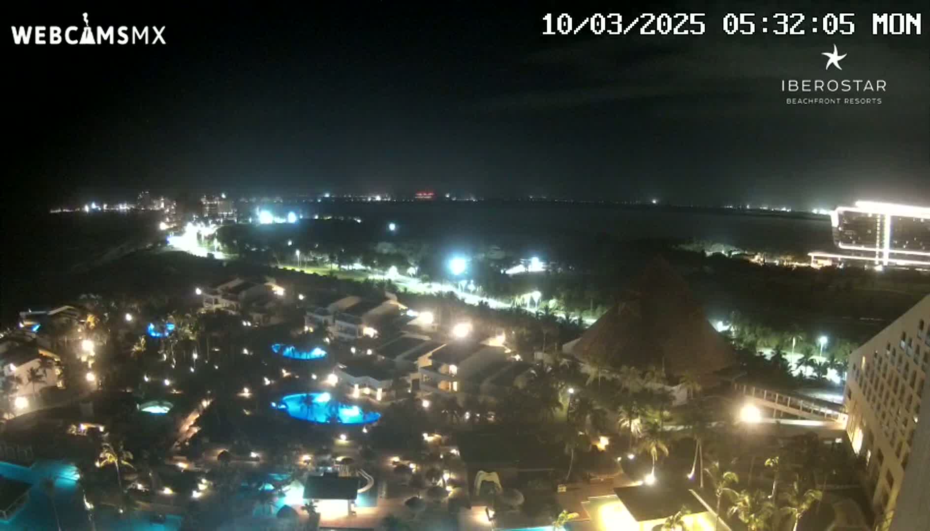 A nighttime, aerial view of a resort complex with illuminated buildings, pools, and landscaping, overlooking a dark body of water.