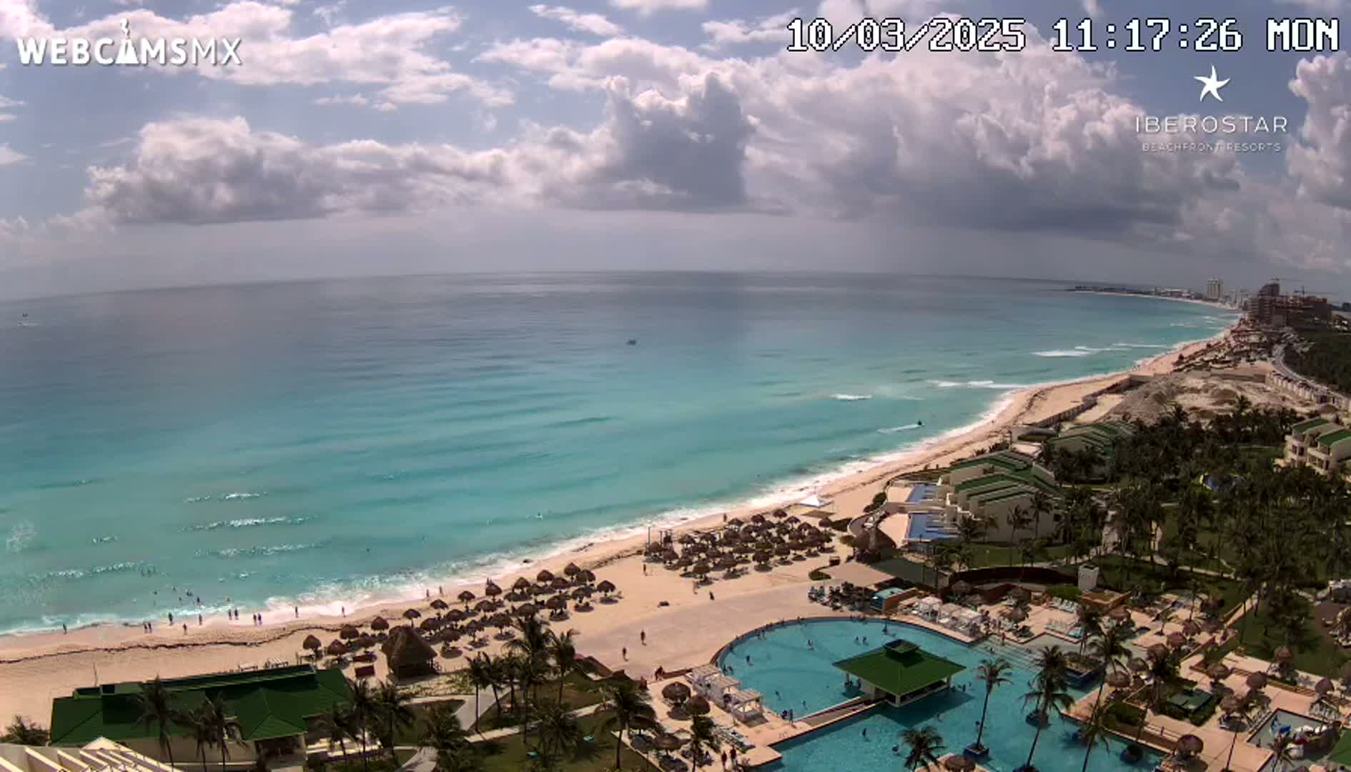 An aerial view shows a resort with multiple pools overlooking a beach with turquoise water under a partly cloudy sky.