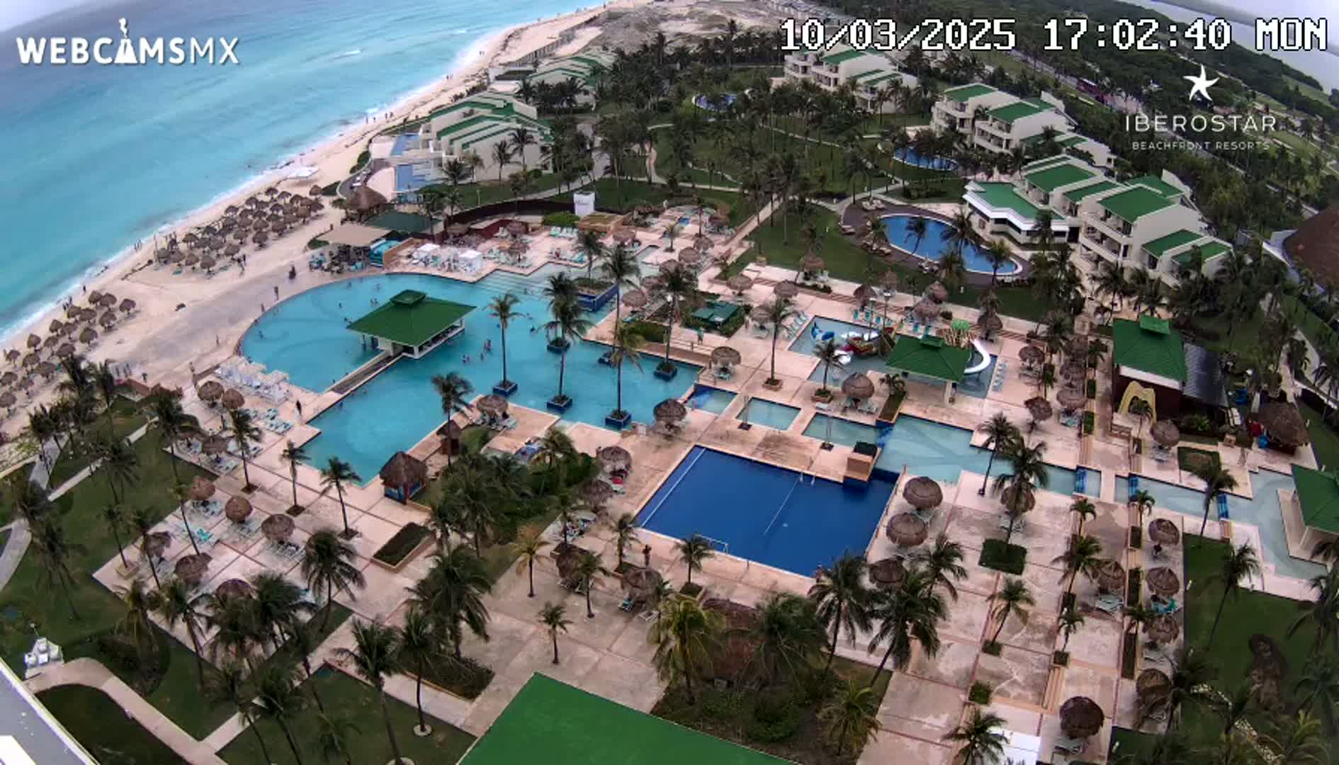 An aerial view shows a resort with numerous pools, palm trees, and a beach under partly cloudy skies.