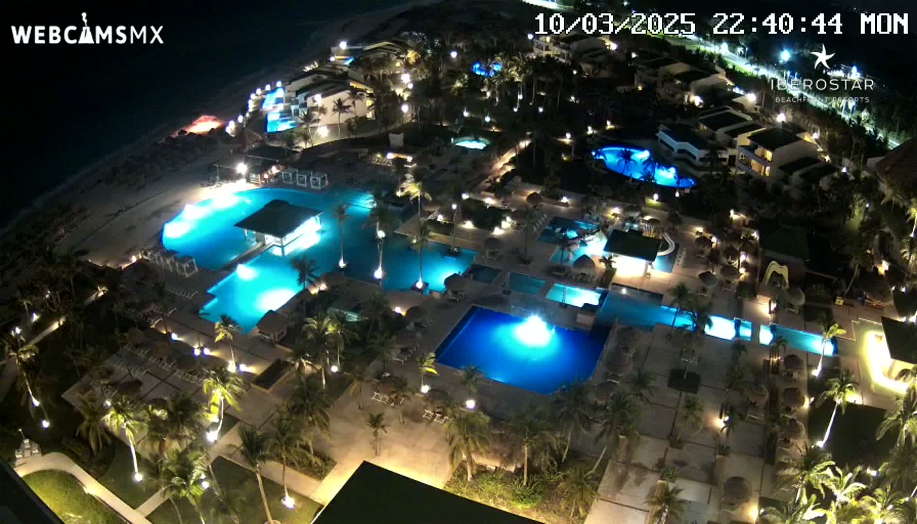 An aerial nighttime view of a resort with numerous illuminated swimming pools surrounded by palm trees and buildings.