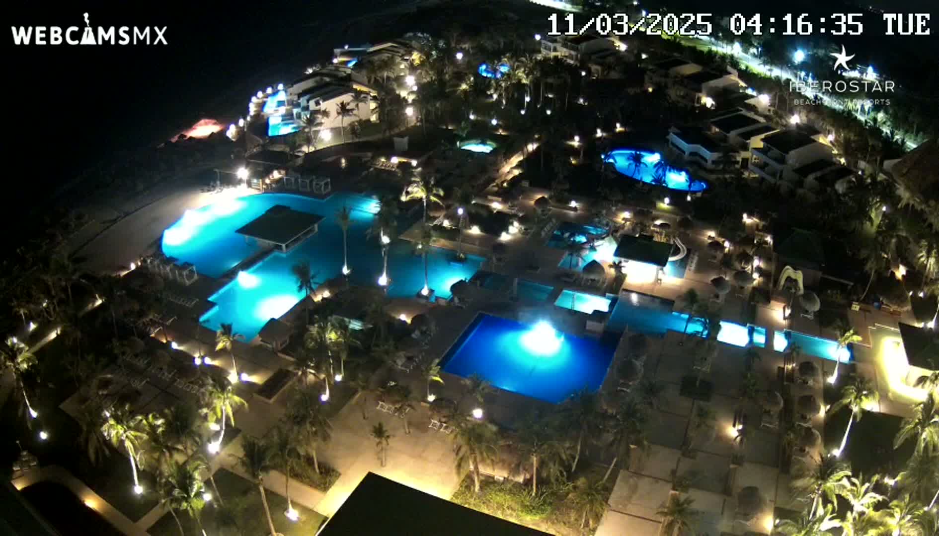 A nighttime aerial view of a resort complex with numerous illuminated swimming pools surrounded by palm trees and buildings.
