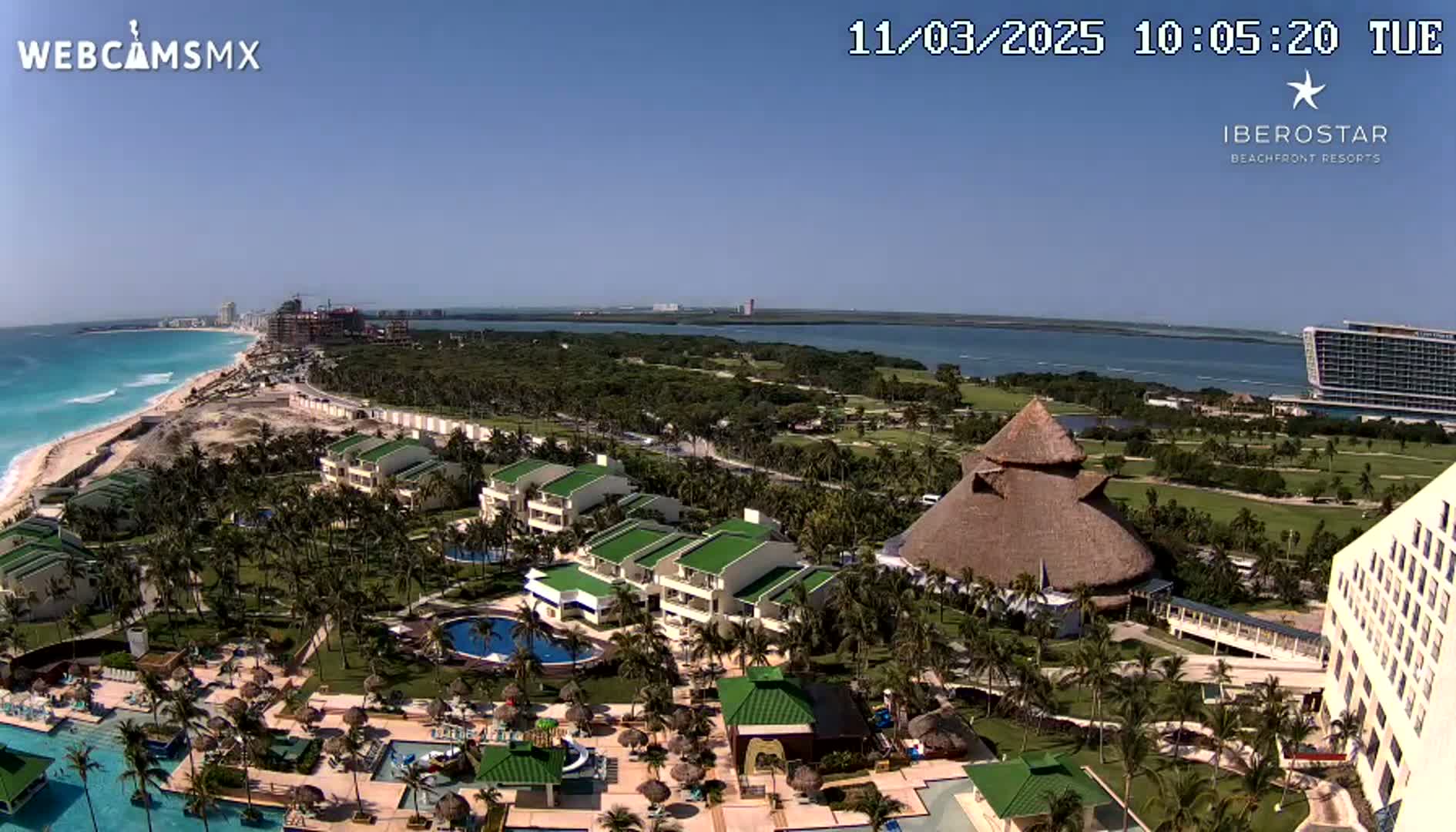 An aerial view of a resort complex on a sunny day, showing buildings, pools, palm trees, and a beach with turquoise water in the distance.