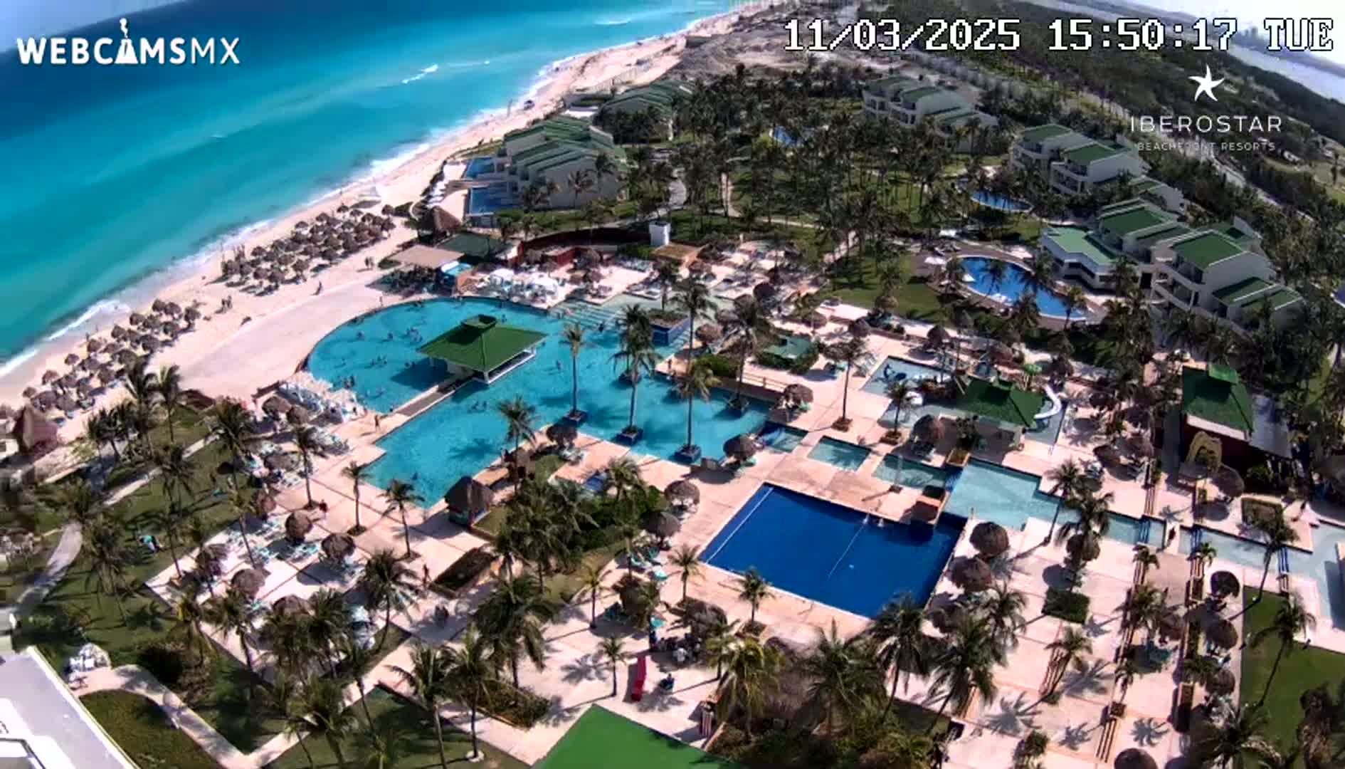 An aerial view shows a beach resort with multiple pools, palm trees, and a sandy beach next to a turquoise ocean on a sunny day.