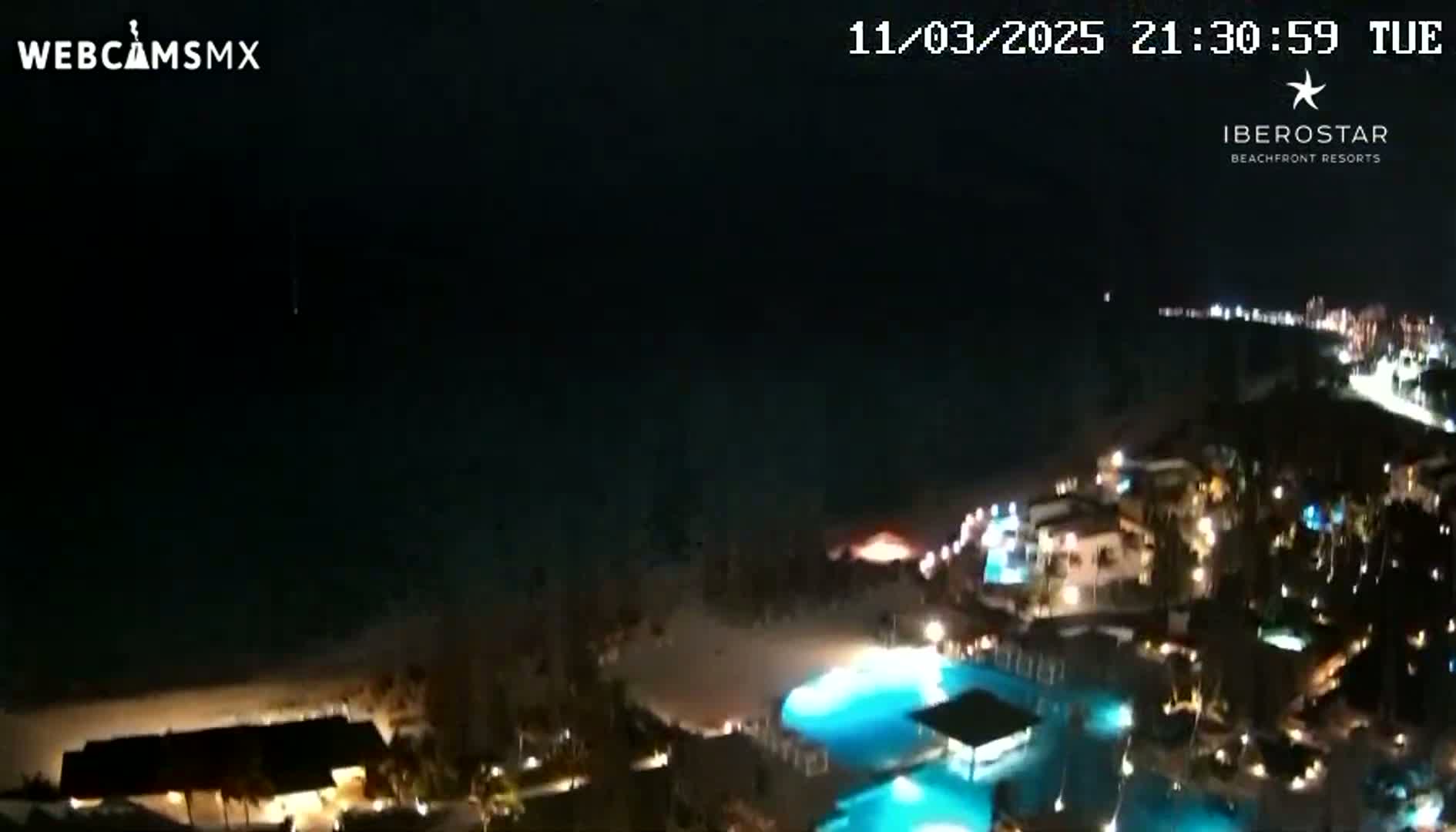 A nighttime aerial view shows a resort area along a beach, with illuminated pools and buildings under a clear, dark sky.