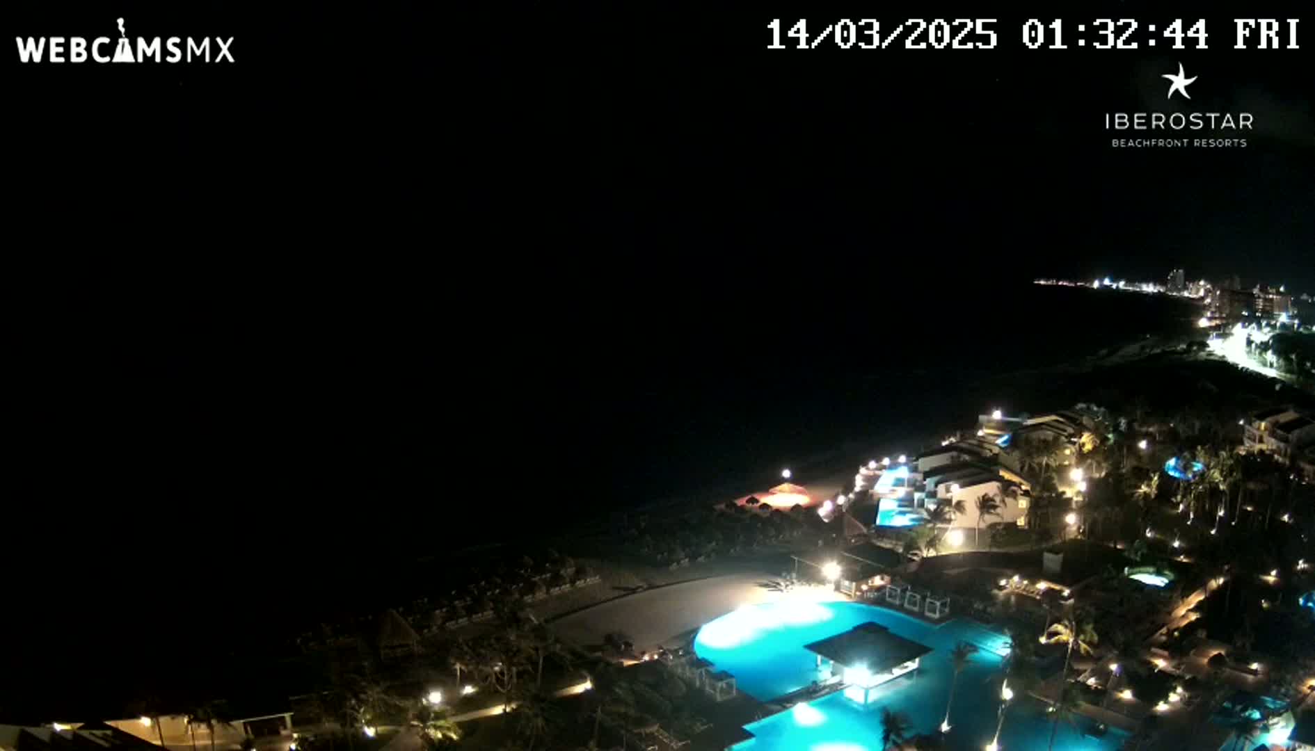 A nighttime aerial view of a resort complex situated on a coastline, featuring illuminated pools and buildings under a dark sky.