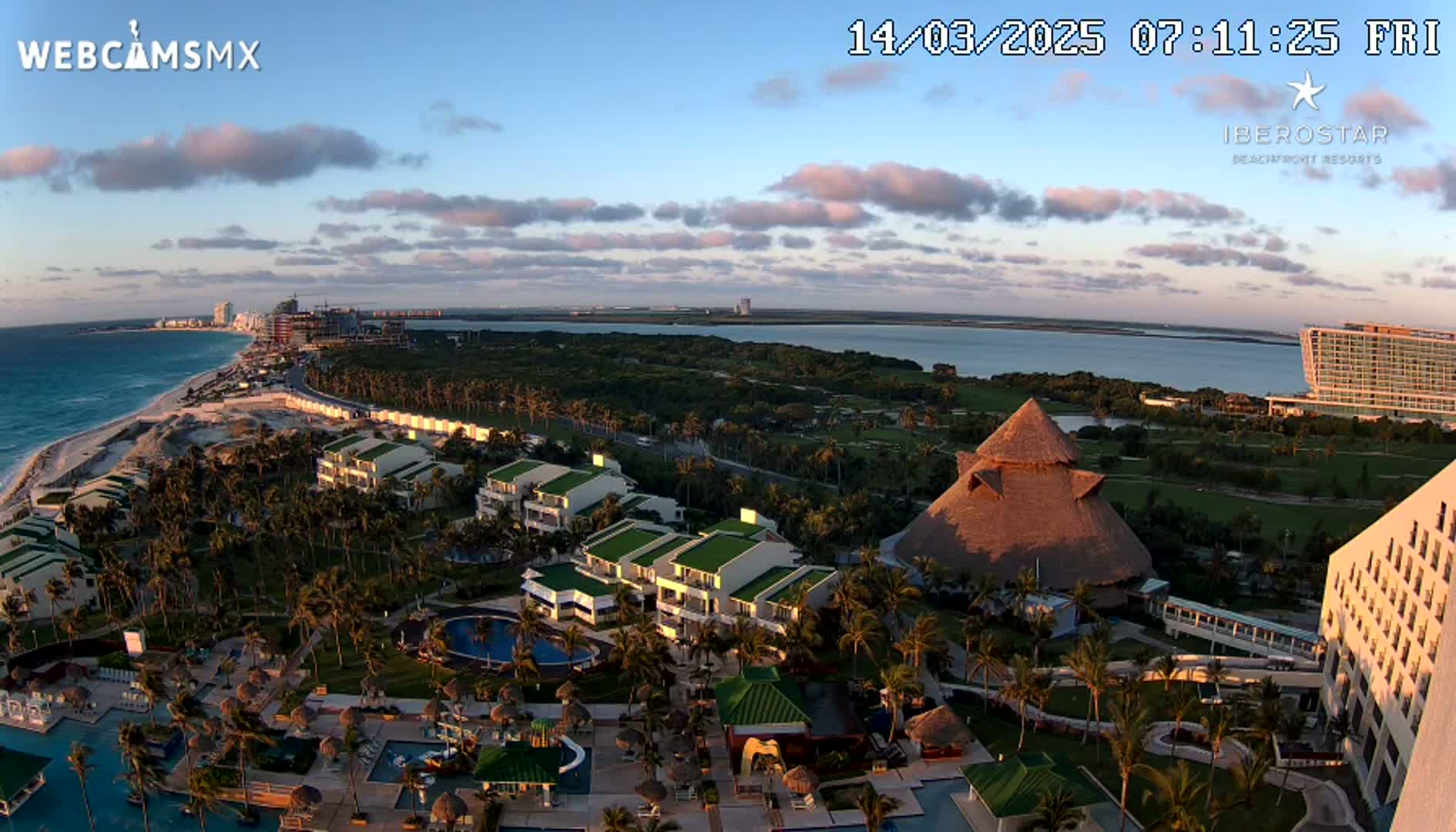 An aerial view of a tropical resort at sunrise, showing buildings, pools, palm trees, and a beach with clear blue water under partly cloudy skies.