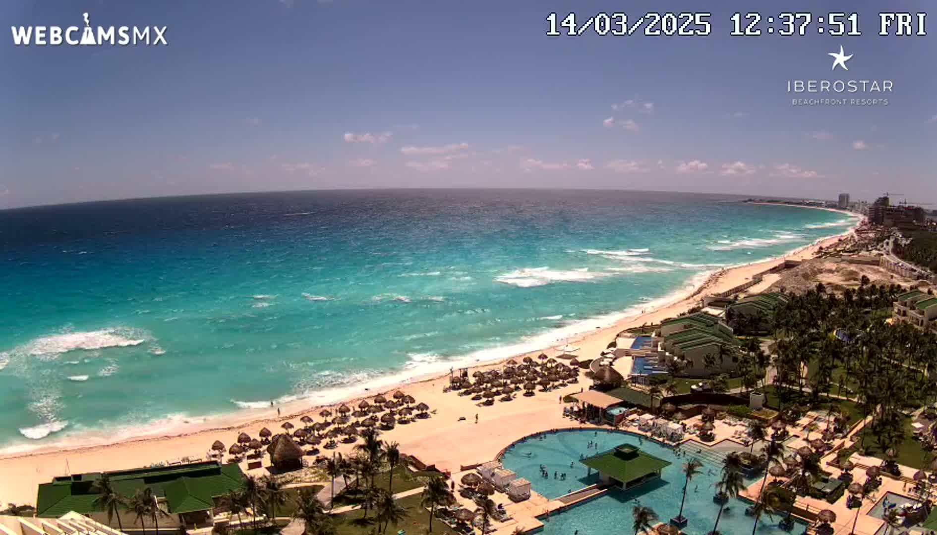 An aerial view shows a resort on a sandy beach with turquoise water and waves under a mostly sunny sky.