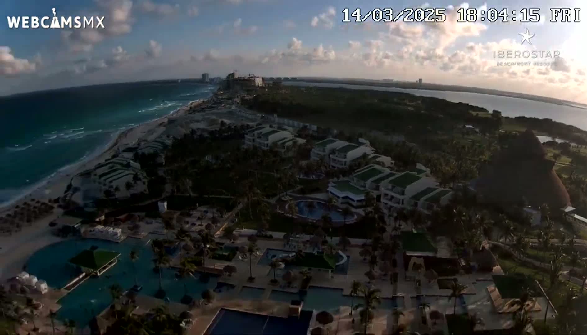 An aerial view shows a resort complex on a beach with turquoise water under a partly cloudy sky.