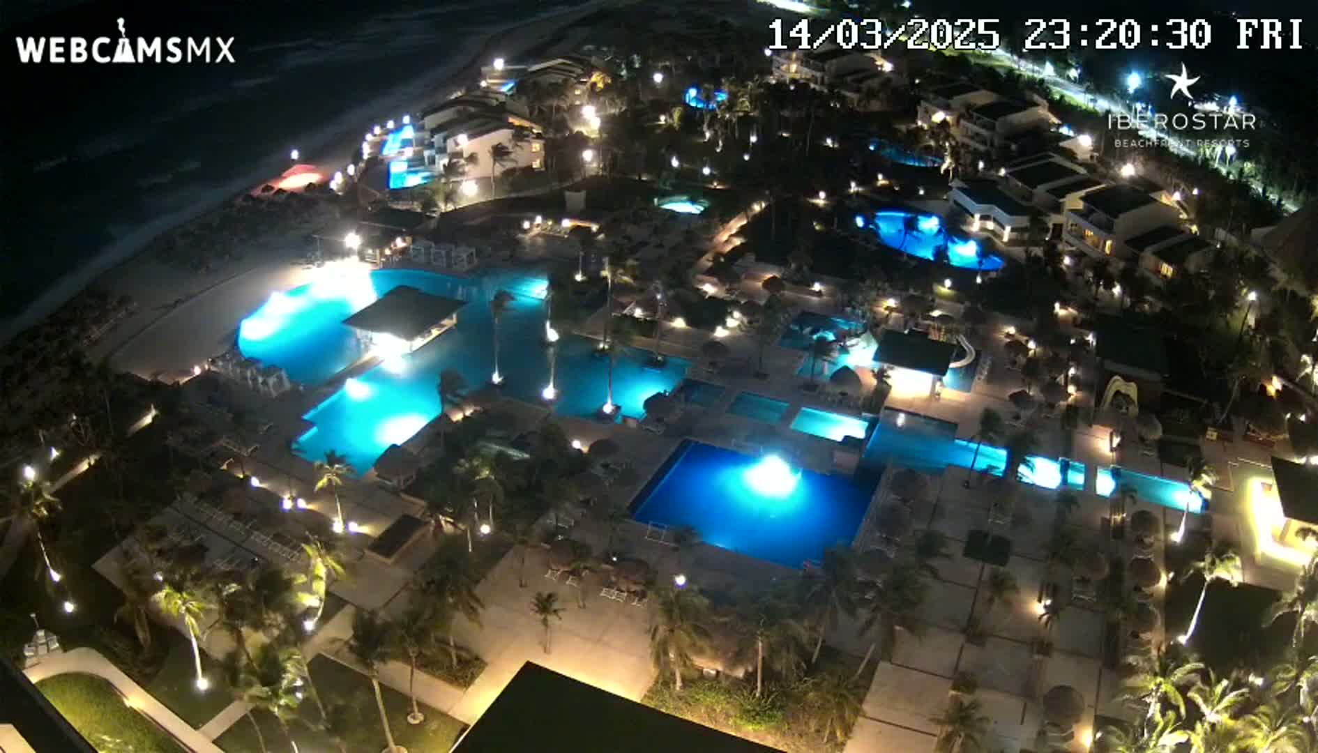 A nighttime aerial view of a resort complex with numerous illuminated swimming pools, surrounding buildings, and palm trees.