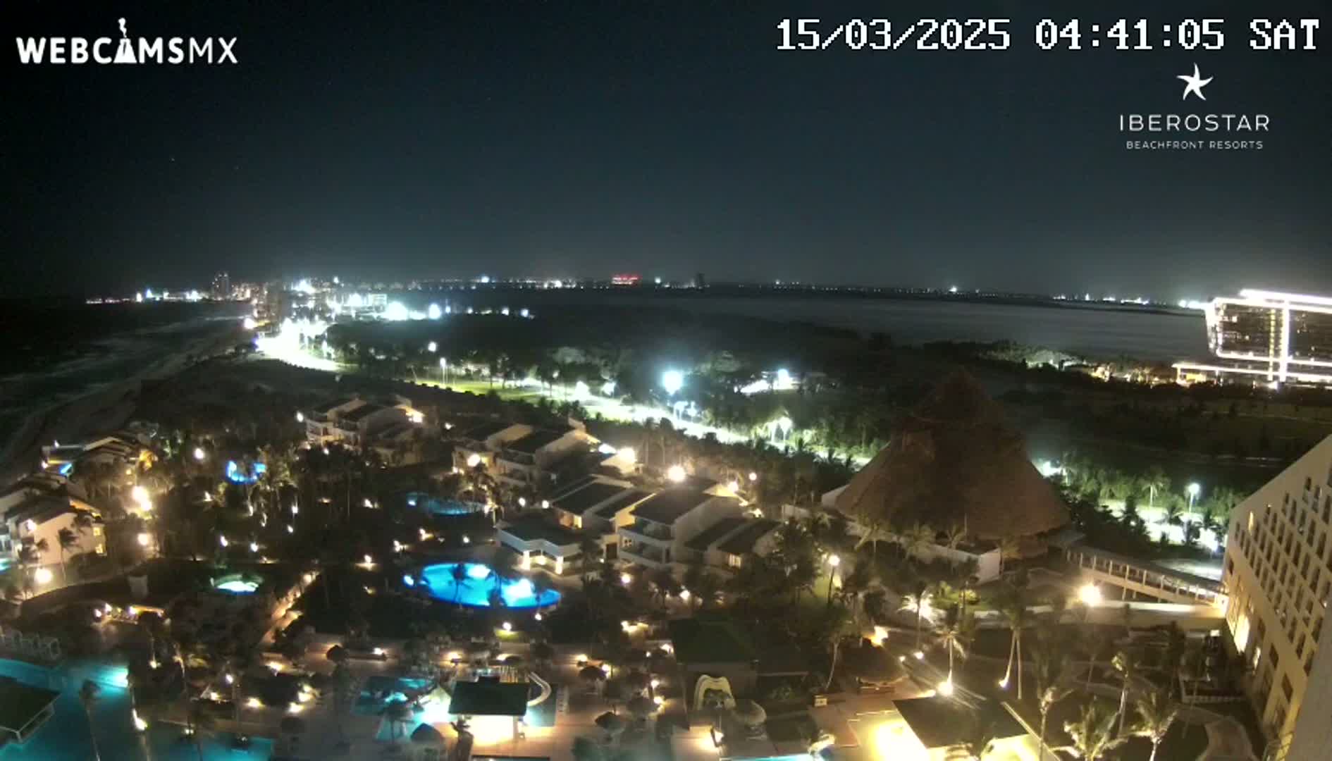 A nighttime, aerial view of a resort complex with illuminated buildings, pools, and landscaping, near a body of water and a city skyline under a clear sky.