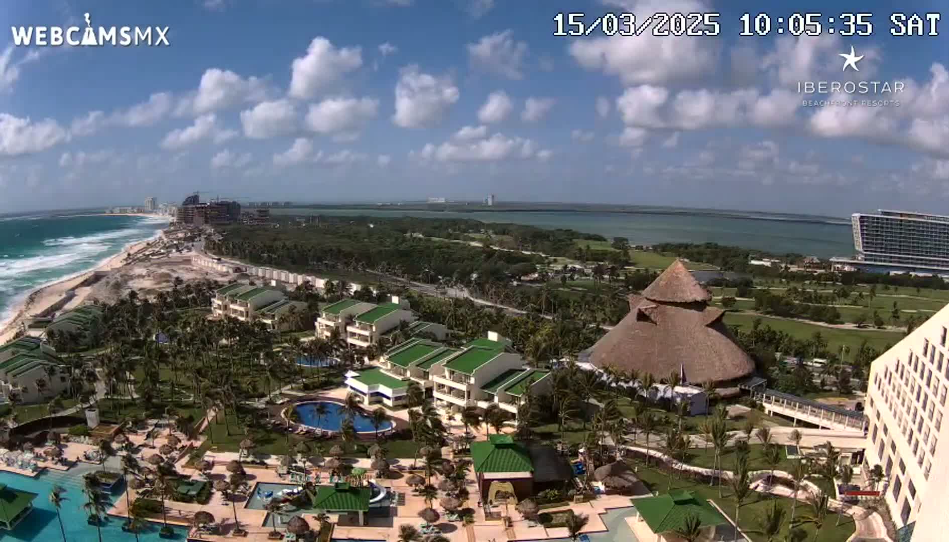 An aerial view of a tropical resort on a partly cloudy day, showing buildings, pools, palm trees, and a beach with ocean waves.