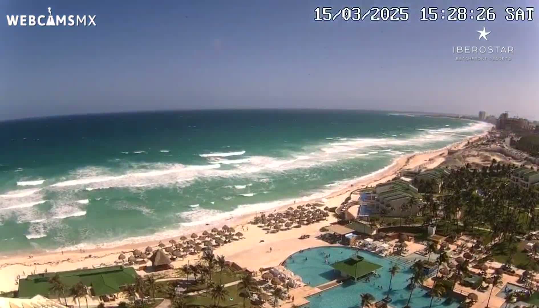 A beach resort with multiple pools overlooks a turquoise ocean with moderate waves under a clear, sunny sky.