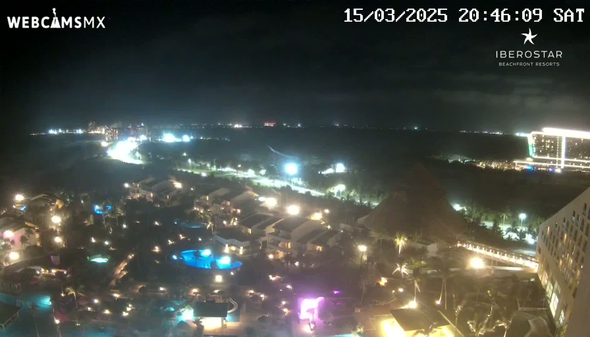 A nighttime aerial view of a resort complex with illuminated buildings, pools, and landscaping, overlooking a dark ocean under a clear sky.