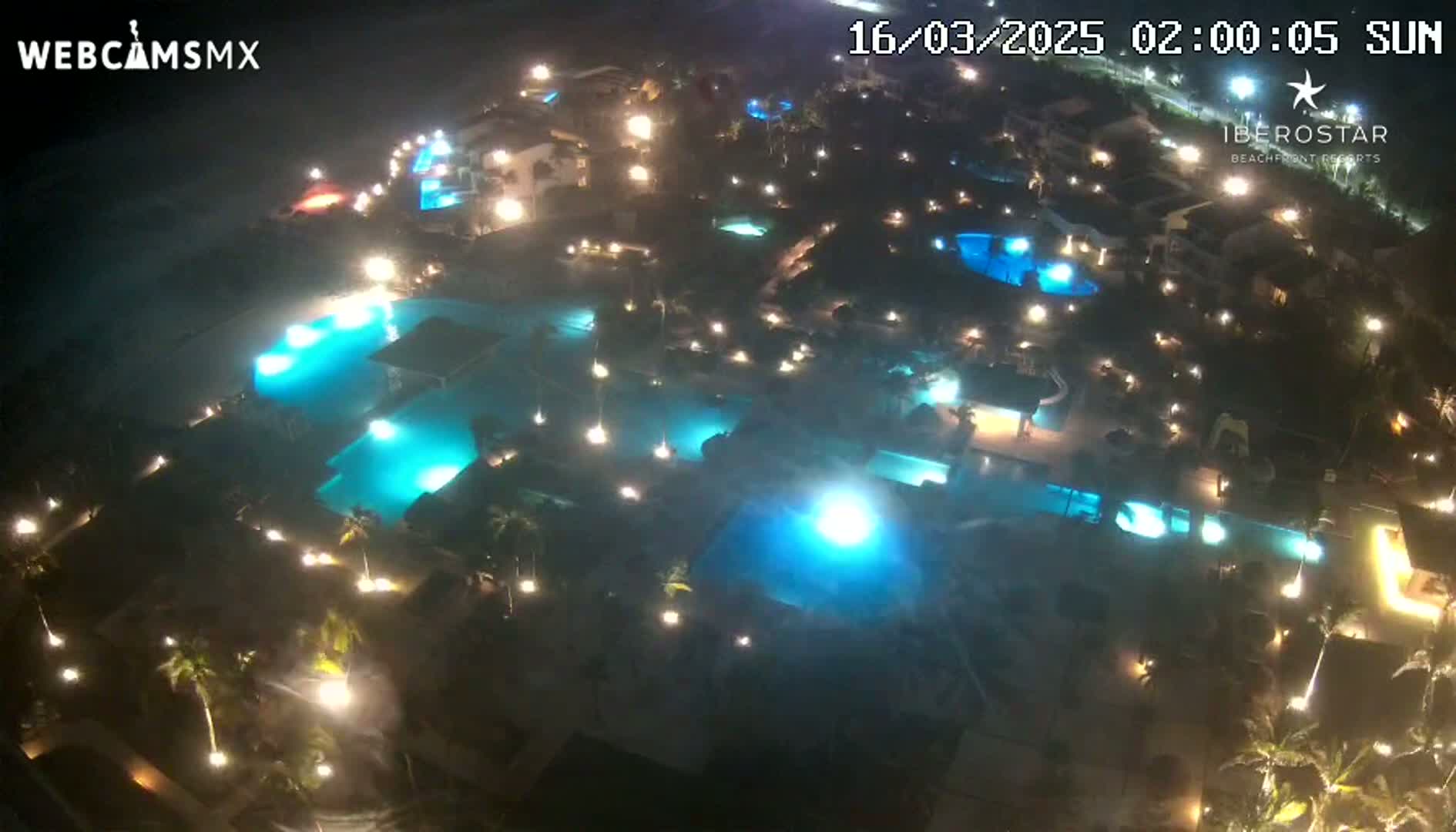 An aerial nighttime view shows a resort complex with numerous illuminated swimming pools and surrounding landscaping.